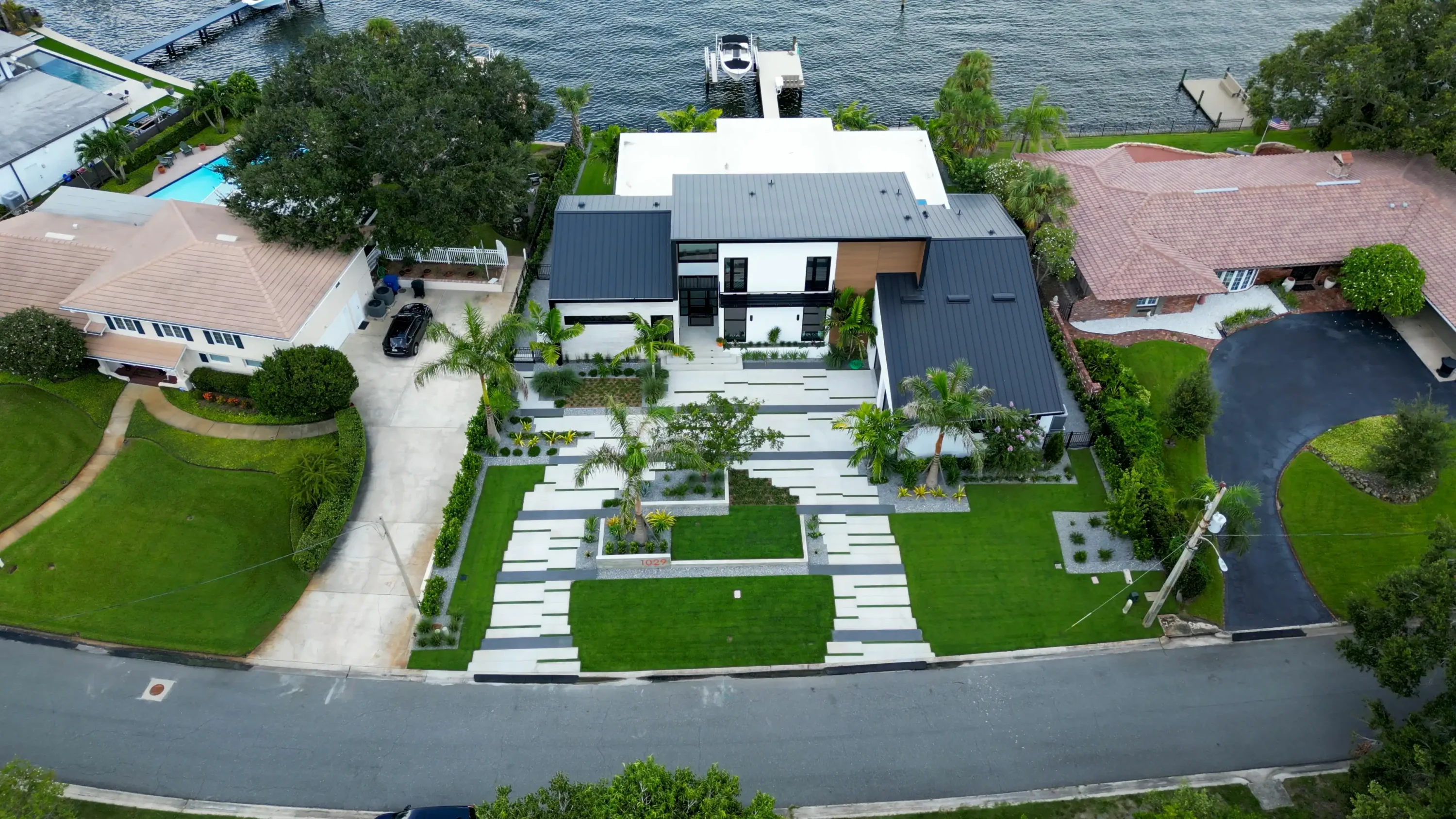 Aerial view of contemporary white and black home with geometric lawn patterns, palm trees, and boat dock on Florida waterfront.