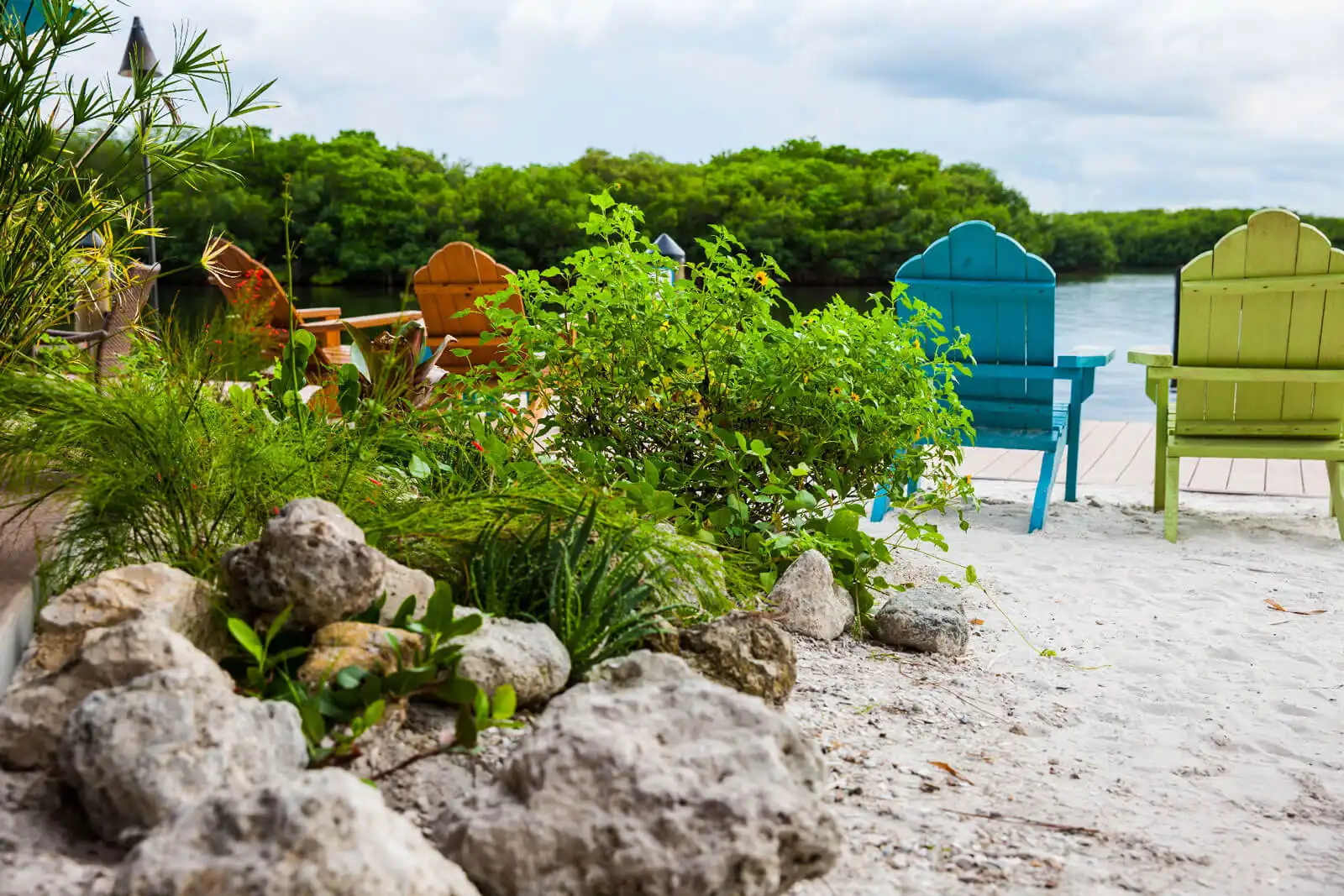 Blue and green Adirondack chairs on sandy beach overlooking calm water, with lush mangroves and tropical vegetation in St Petersburg, FL luxury landscape design.