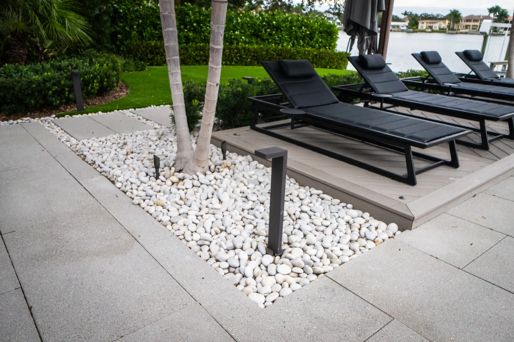 Luxury waterfront patio with black loungers, white river rock landscaping, and palm trees overlooking water in St Petersburg, FL.