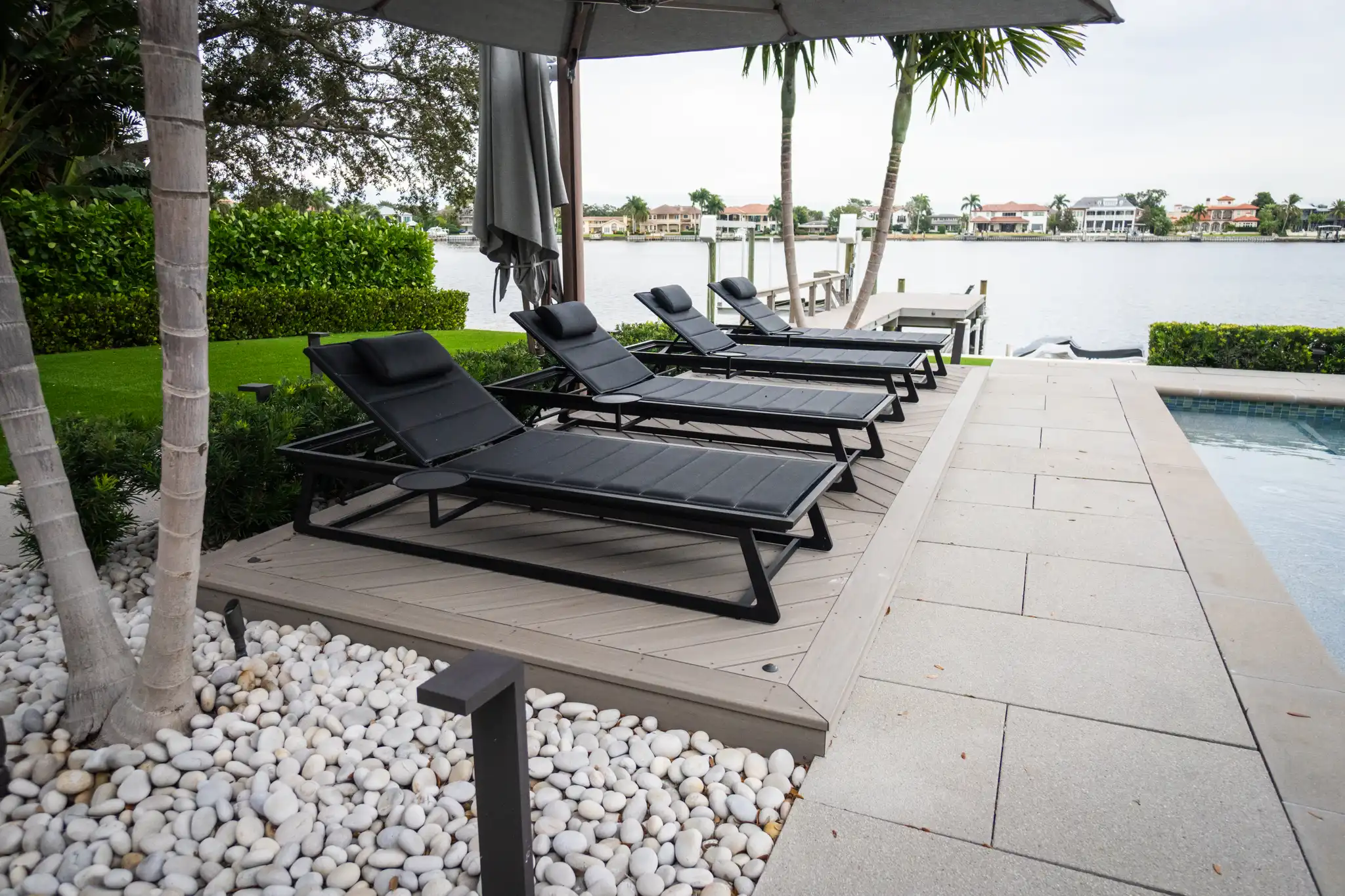 Luxury waterfront pool deck with black loungers under gray umbrella, manicured lawn, and residential canal homes across the water in St Petersburg, FL.