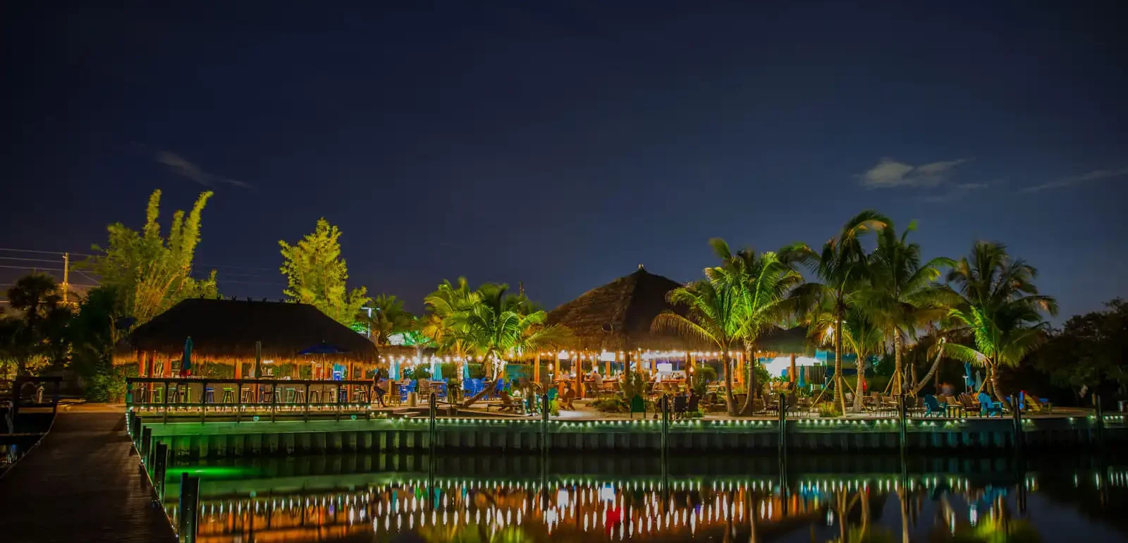 Illuminated luxury waterfront resort with tiki-style architecture, palm trees, and green dock reflecting in calm water at dusk in St Petersburg.