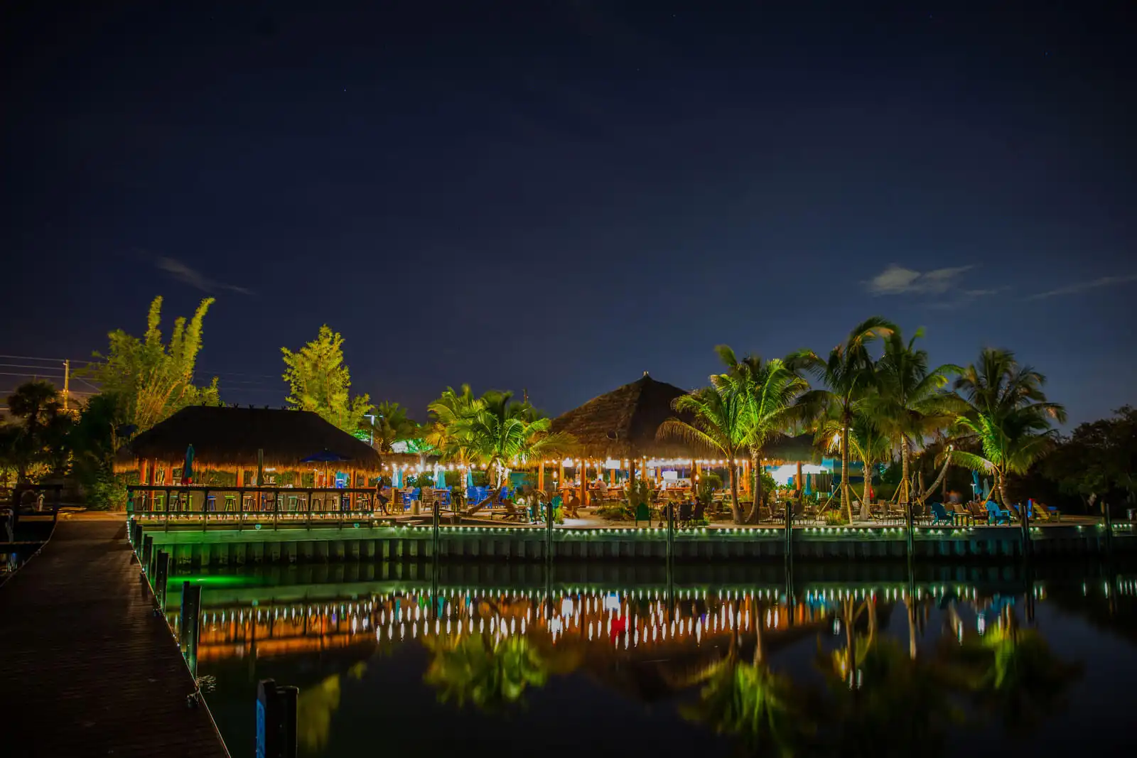 Illuminated luxury waterfront resort with tiki-style architecture, palm trees, and green dock reflecting in calm water at dusk in St Petersburg.