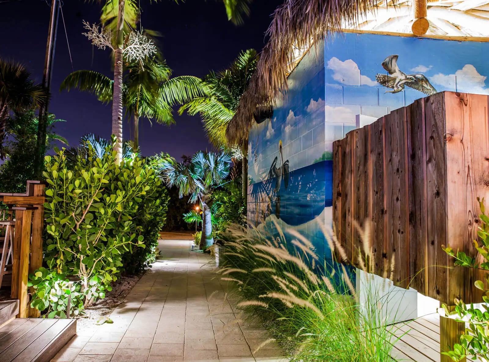 Night view of luxurious courtyard featuring ocean mural, wooden fence, and lush tropical landscaping with palm trees and coastal design elements.