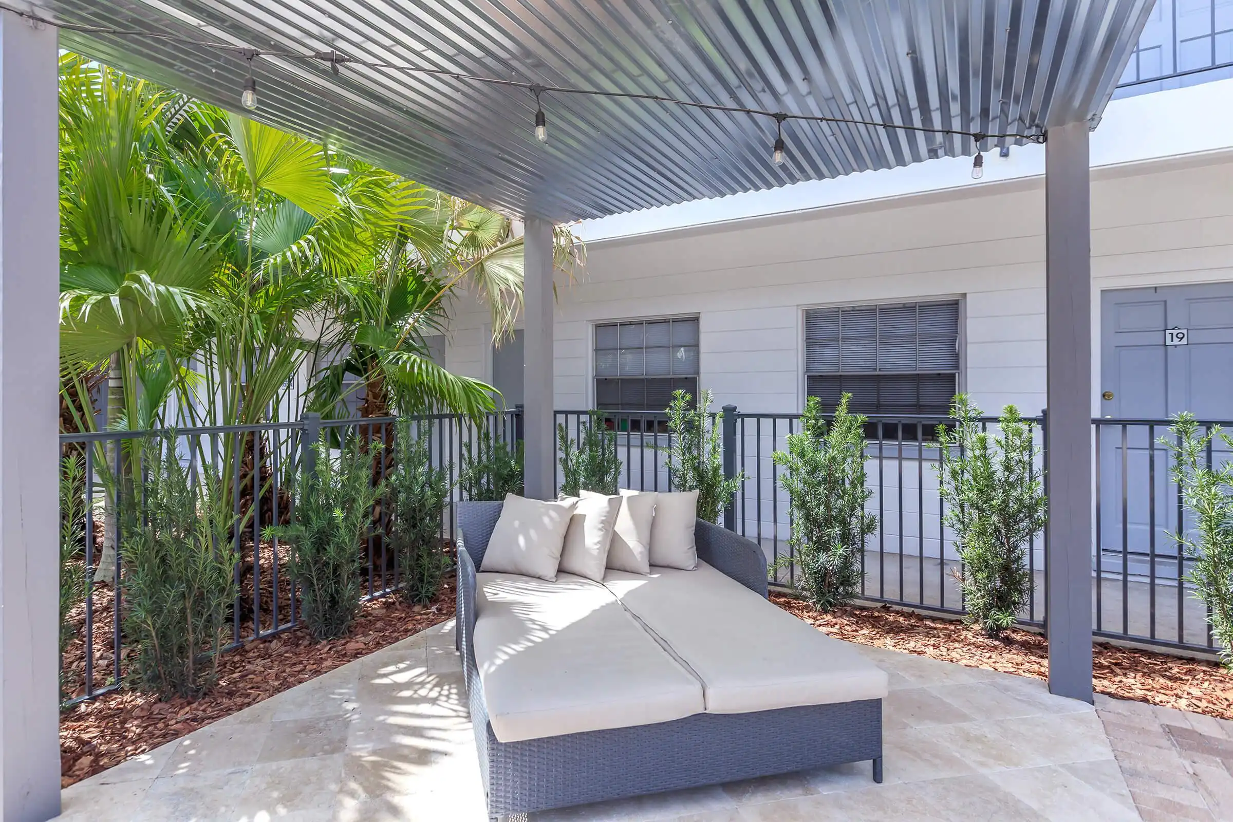 Luxury outdoor patio featuring a gray wicker daybed with cream cushions beneath a corrugated metal pergola, flanked by palm trees and architectural plants.
