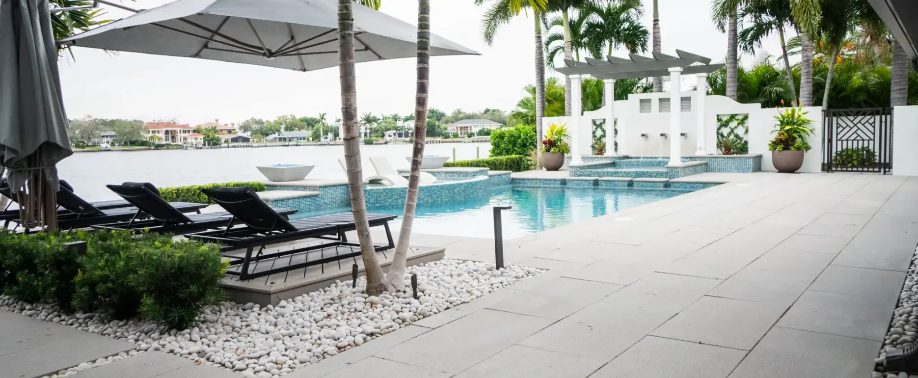 Luxury poolside terrace with gray umbrellas, black loungers, and palm trees overlooking a waterfront in St Petersburg, Florida, featuring modern pool design and landscape architecture.