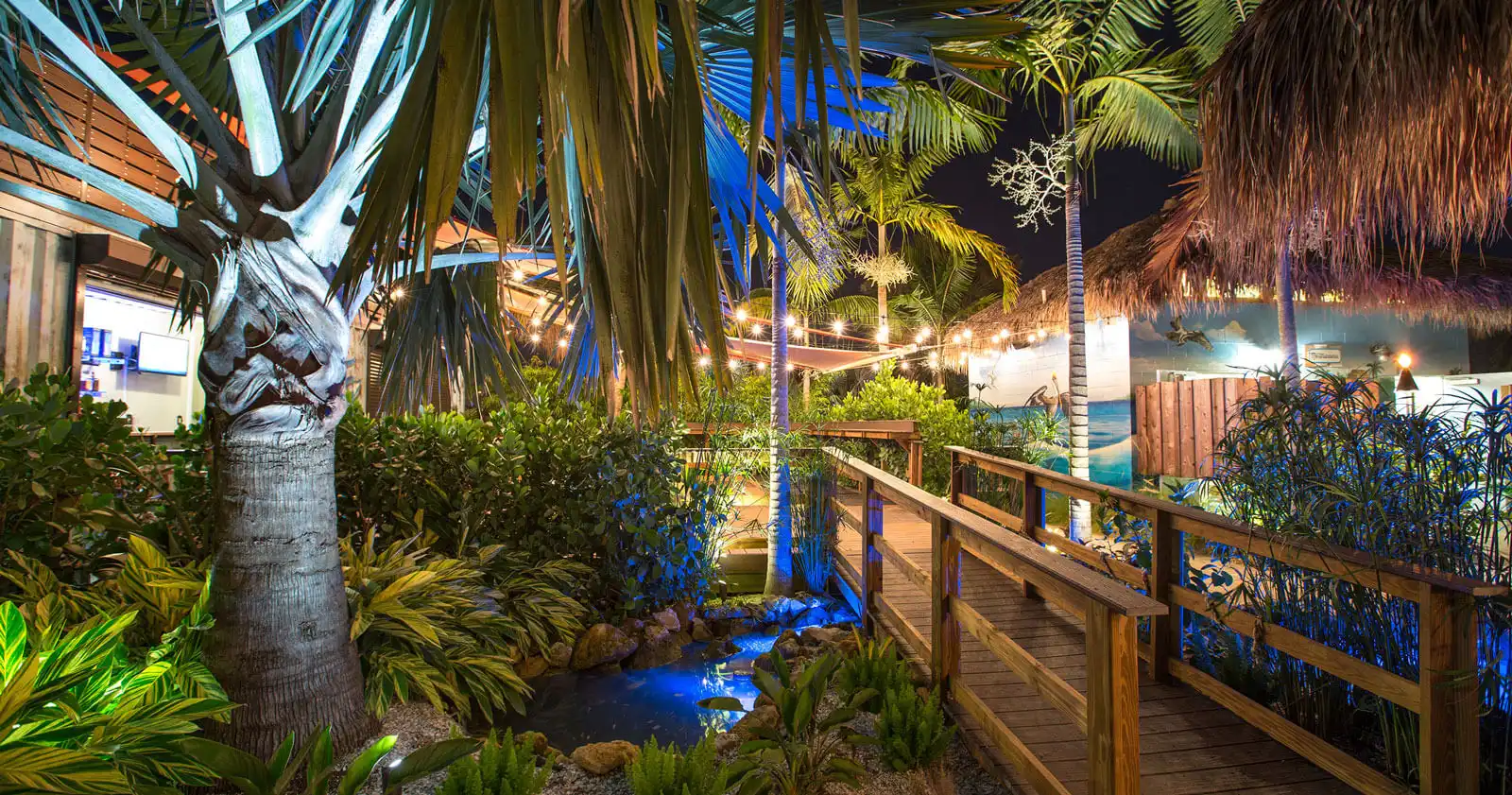 Illuminated wooden boardwalk lined with palm trees and lush landscaping at night, featuring blue accent lighting and tropical vegetation in a St Petersburg FL residential community.