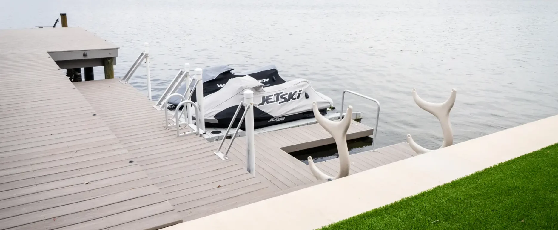 Modern composite dock with white jet ski lift and moored watercraft, luxury St Petersburg residential community across the bay with manicured lawn foreground.
