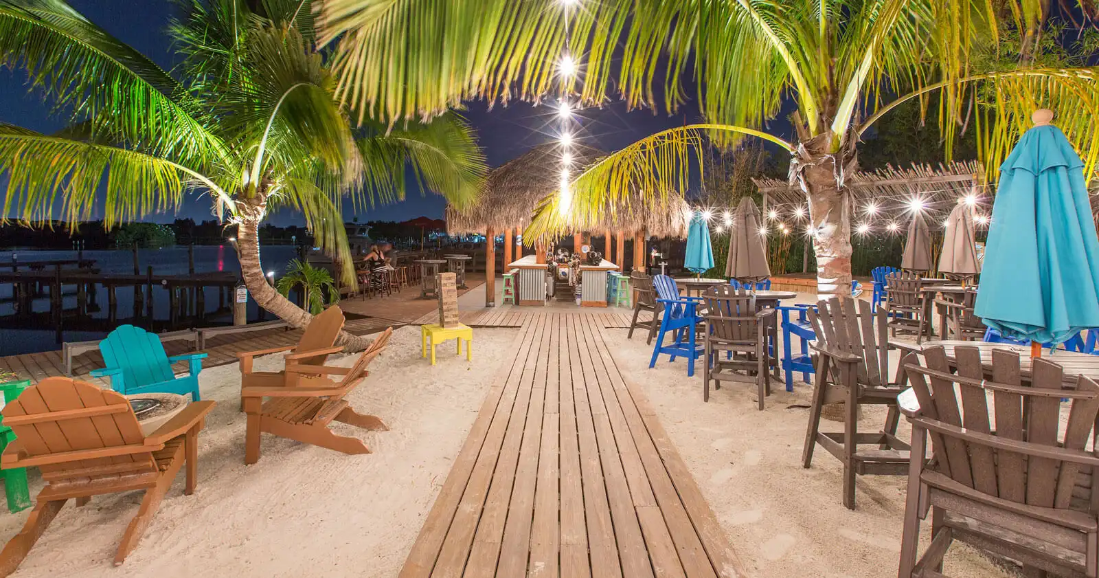 Illuminated beachfront venue with palm-thatched structures, wooden boardwalk, colorful Adirondack chairs, and dining areas overlooking water at night in St. Petersburg, FL.