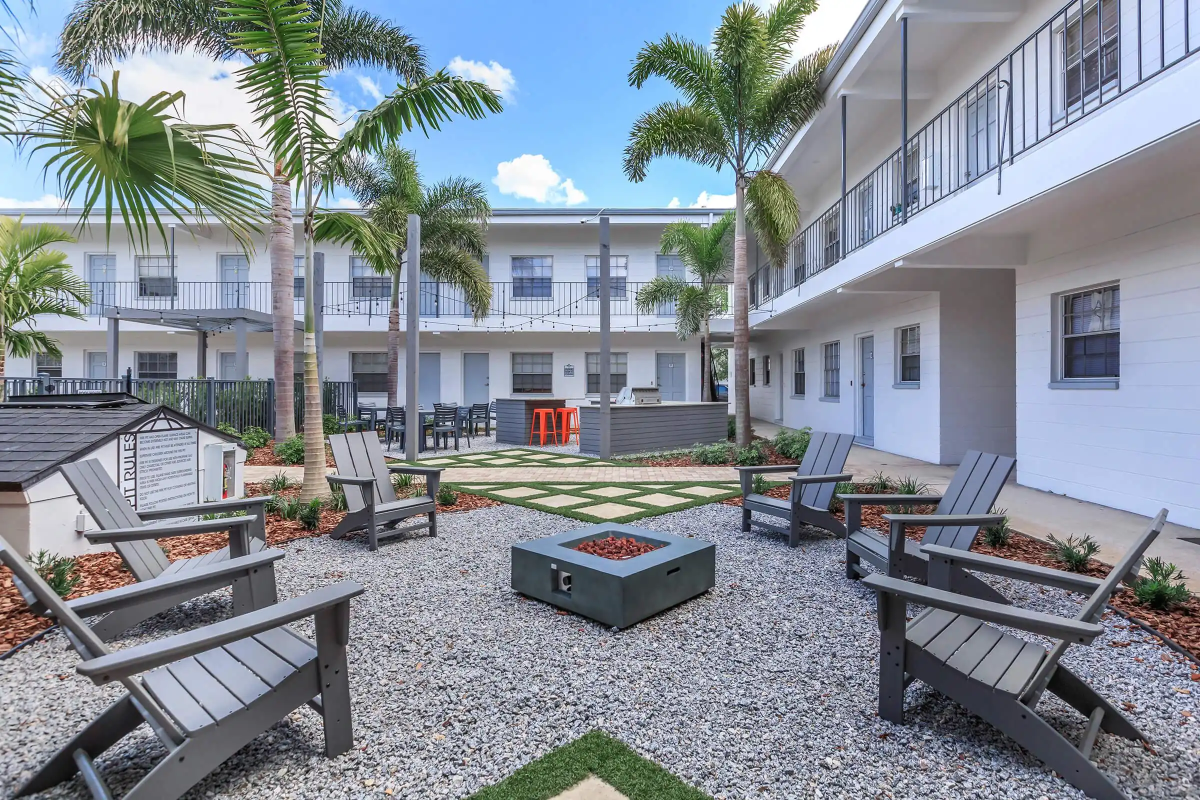 Modern residential courtyard with Adirondack chairs surrounding a central fire pit, framed by white multi-story buildings and palm trees in St Petersburg, FL.
