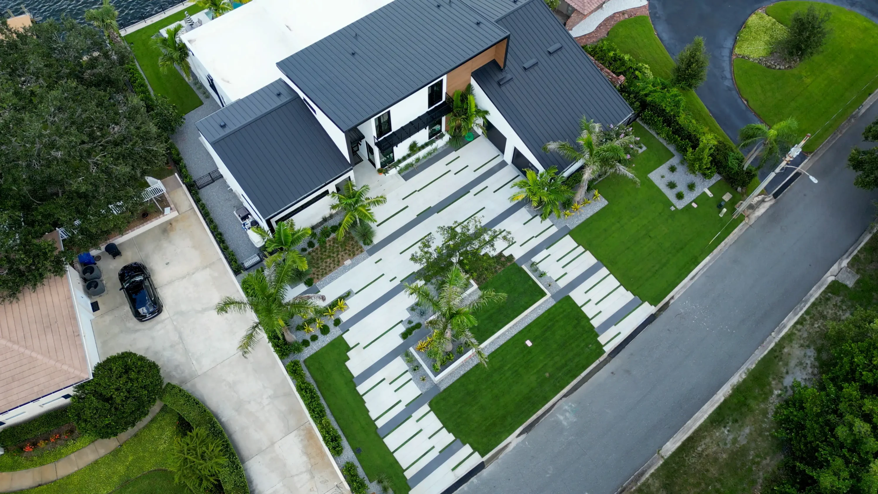 Aerial view of contemporary home with dark metal roof, linear hardscape pathways, palm trees, and manicured lawn in St Petersburg luxury residential community.