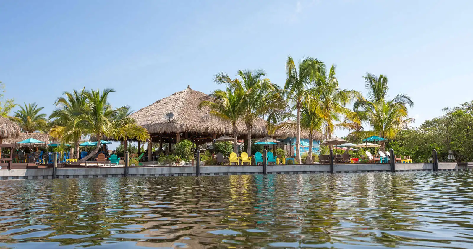 Luxury waterfront resort with thatched-roof pavilion, turquoise umbrellas, and palm trees along St Petersburg waterway.