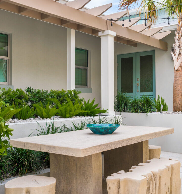 Contemporary outdoor dining area with travertine table and log-slice seating under pergola, featuring tropical landscaping and green modern doors.