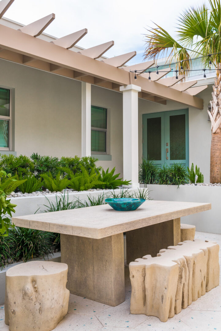Contemporary outdoor dining area with travertine table and log-slice seating under pergola, featuring tropical landscaping and green modern doors.