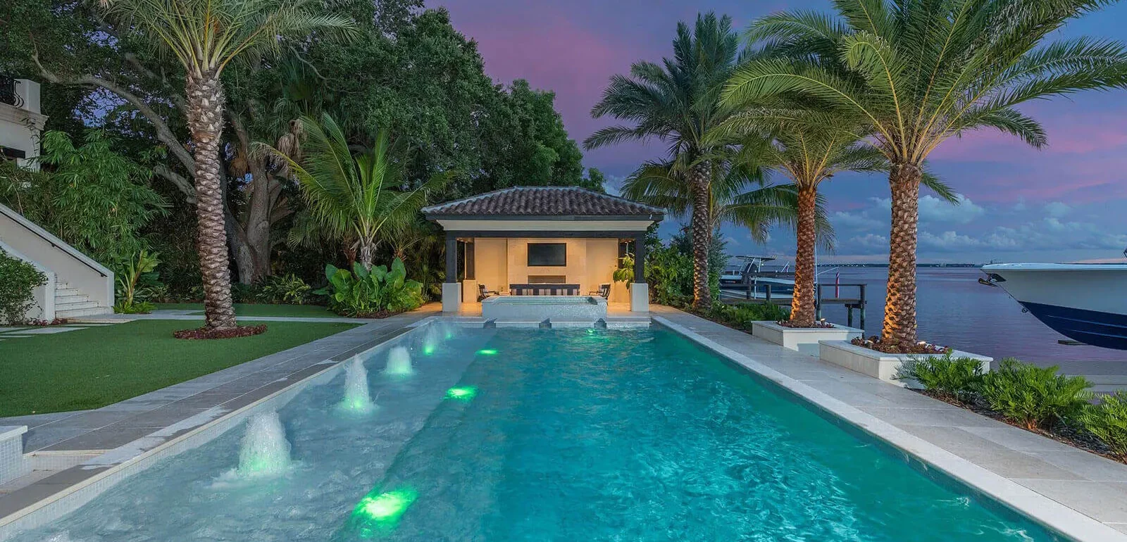 Luxury waterfront pool with green LED lighting, yellow cabana, palm trees, and bay view at dusk in St Petersburg, Florida.