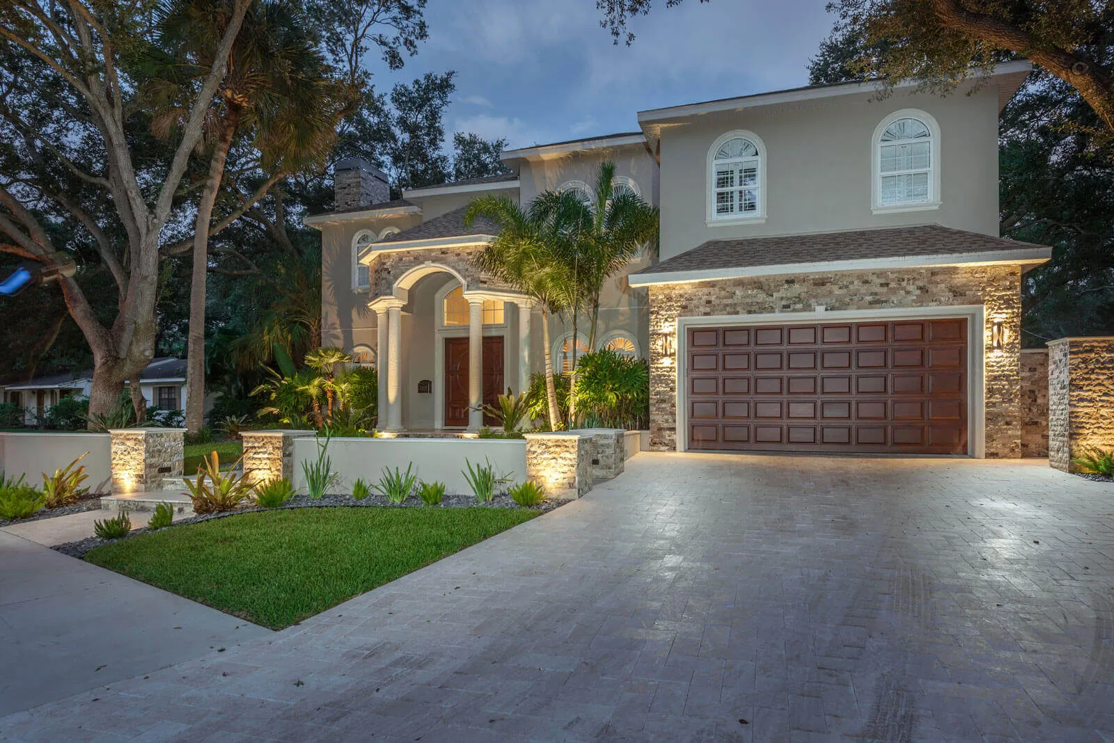 Luxury contemporary home in St Petersburg FL featuring stone facade, arched entryway, and wood grain garage door with professional landscape architecture and ambient lighting.