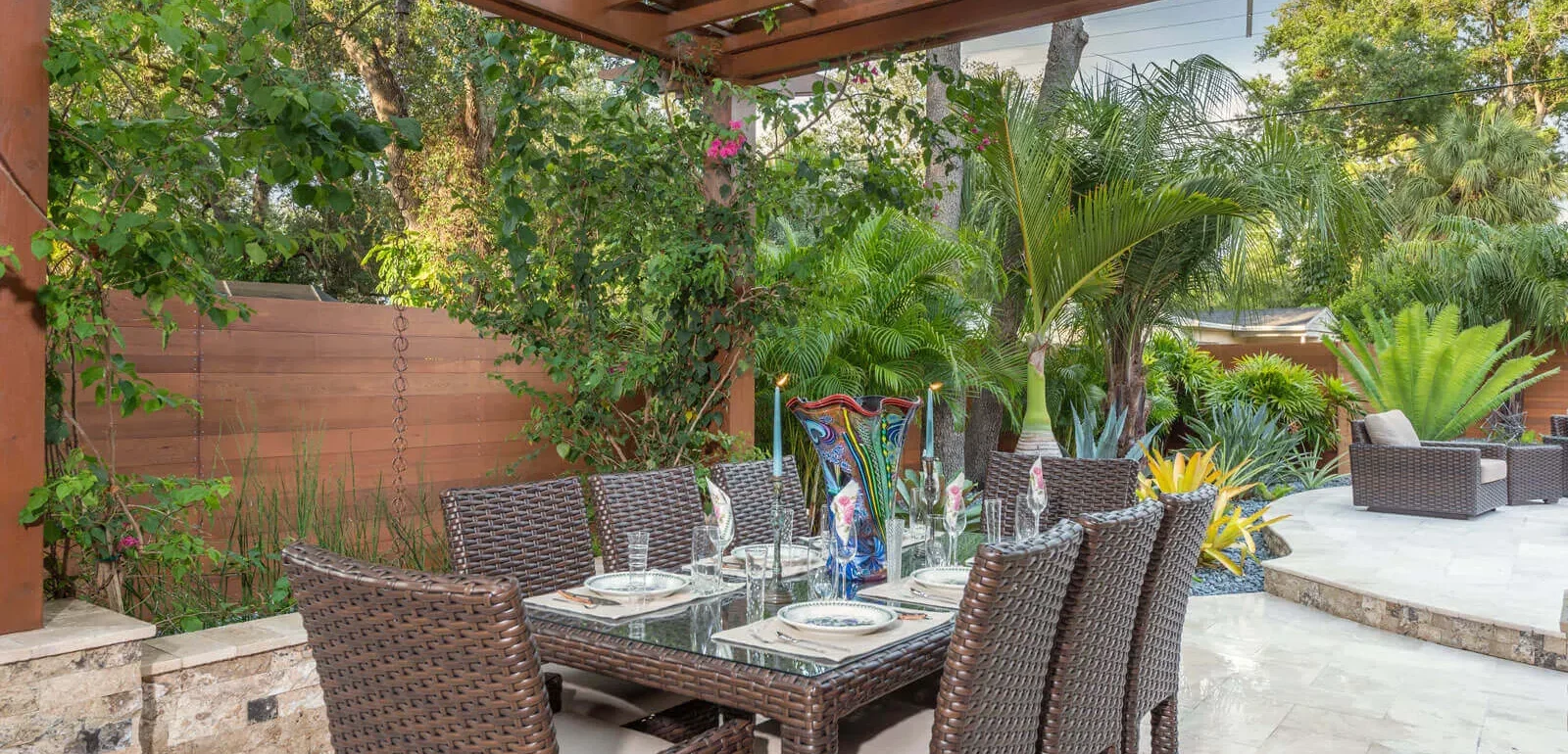 Luxury outdoor dining area with woven rattan furniture beneath wooden pergola, surrounded by tropical landscaping and palms in St Petersburg residential design.