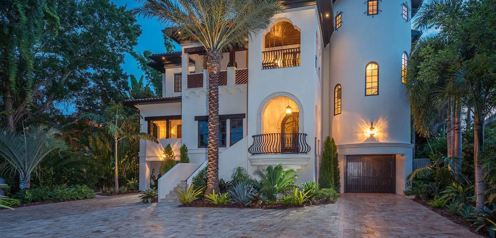 Illuminated white stucco Spanish Colonial mansion with palm trees, arched entryway, and brick driveway in St Petersburg luxury residential community.