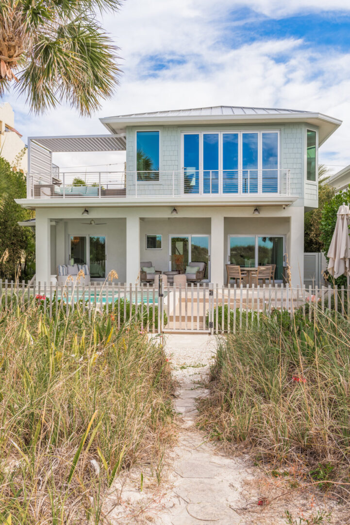 Two-story modern coastal home with white shingle exterior, blue windows, and covered porch overlooking beach grass pathway in St Petersburg, FL.