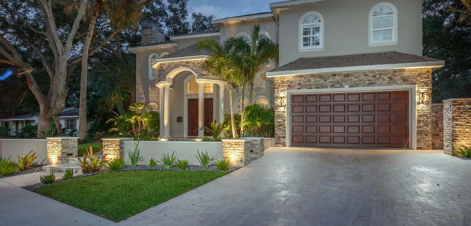 Luxury colonial home with stone facade, arched entry portico, and wood garage doors set on manicured lawn with palm trees in St Petersburg, FL.