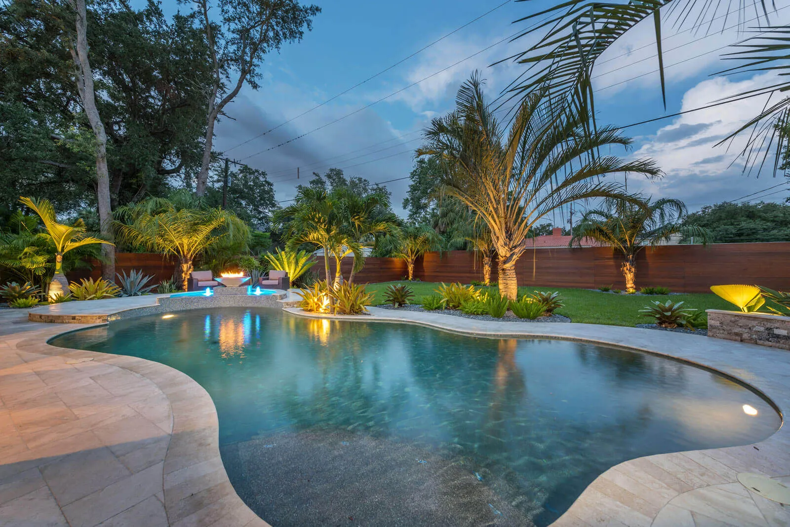 Luxury backyard pool with evening lighting, curved concrete decking, tropical palm trees, and manicured lawn bordered by wooden fence in St Petersburg.