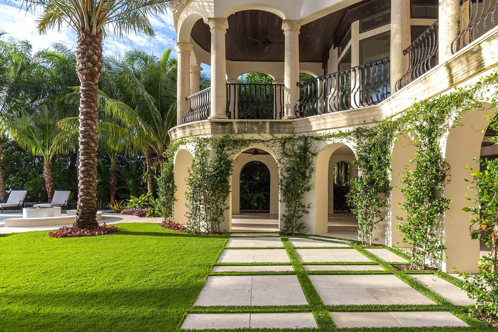Luxury residential courtyard featuring cream stucco architecture with ivy-covered arched alcoves, stepping stone pathway, manicured lawn, and palm trees in St Petersburg.