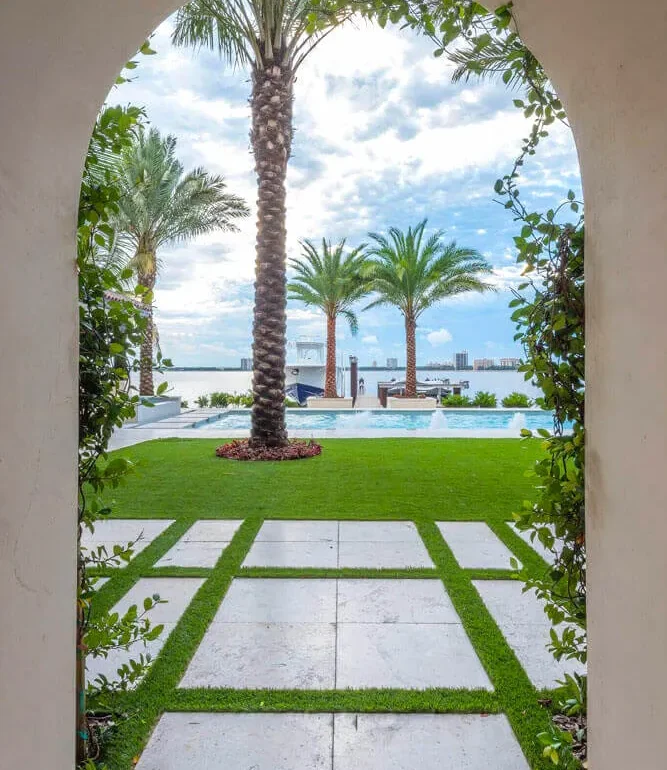 Arched passageway framing manicured lawn, palm trees, and infinity pool overlooking waterfront in St Petersburg luxury residential community.