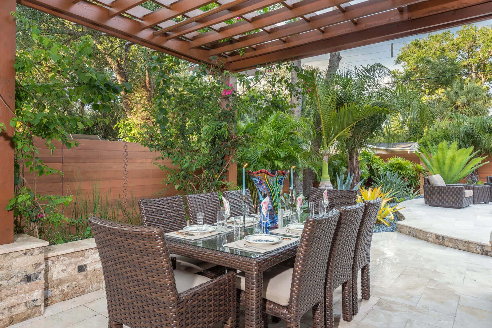 Luxury outdoor dining area with woven rattan chairs and glass-top table under wooden pergola, surrounded by lush tropical plants and palm trees in St Petersburg, FL landscape design.