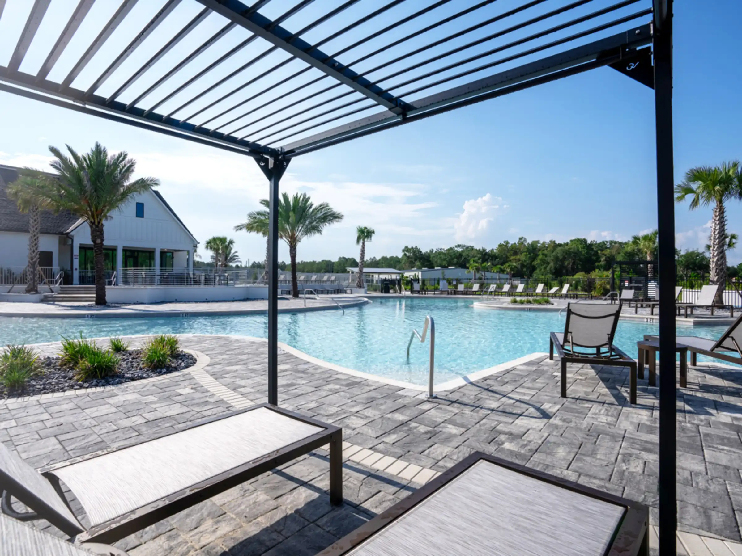 Contemporary pool deck with black pergola shade structure overlooking crystal-blue swimming pool and white amenity building surrounded by palm trees.