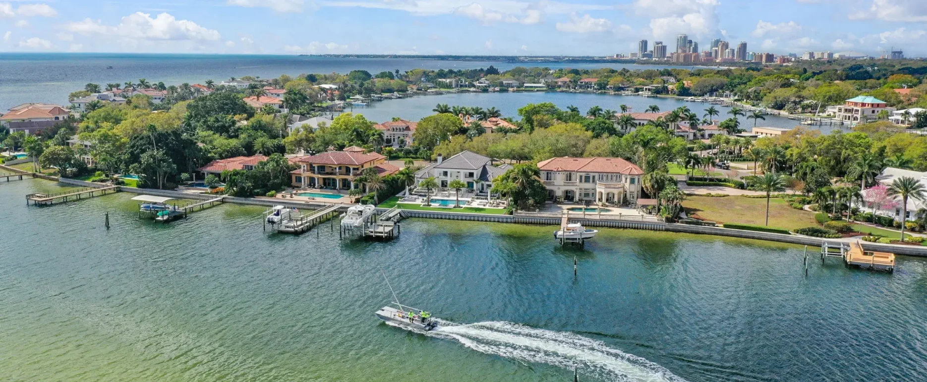 Aerial view of luxury waterfront home with private boat dock, pool, and manicured grounds overlooking Biscayne Bay with Miami skyline in distance.
