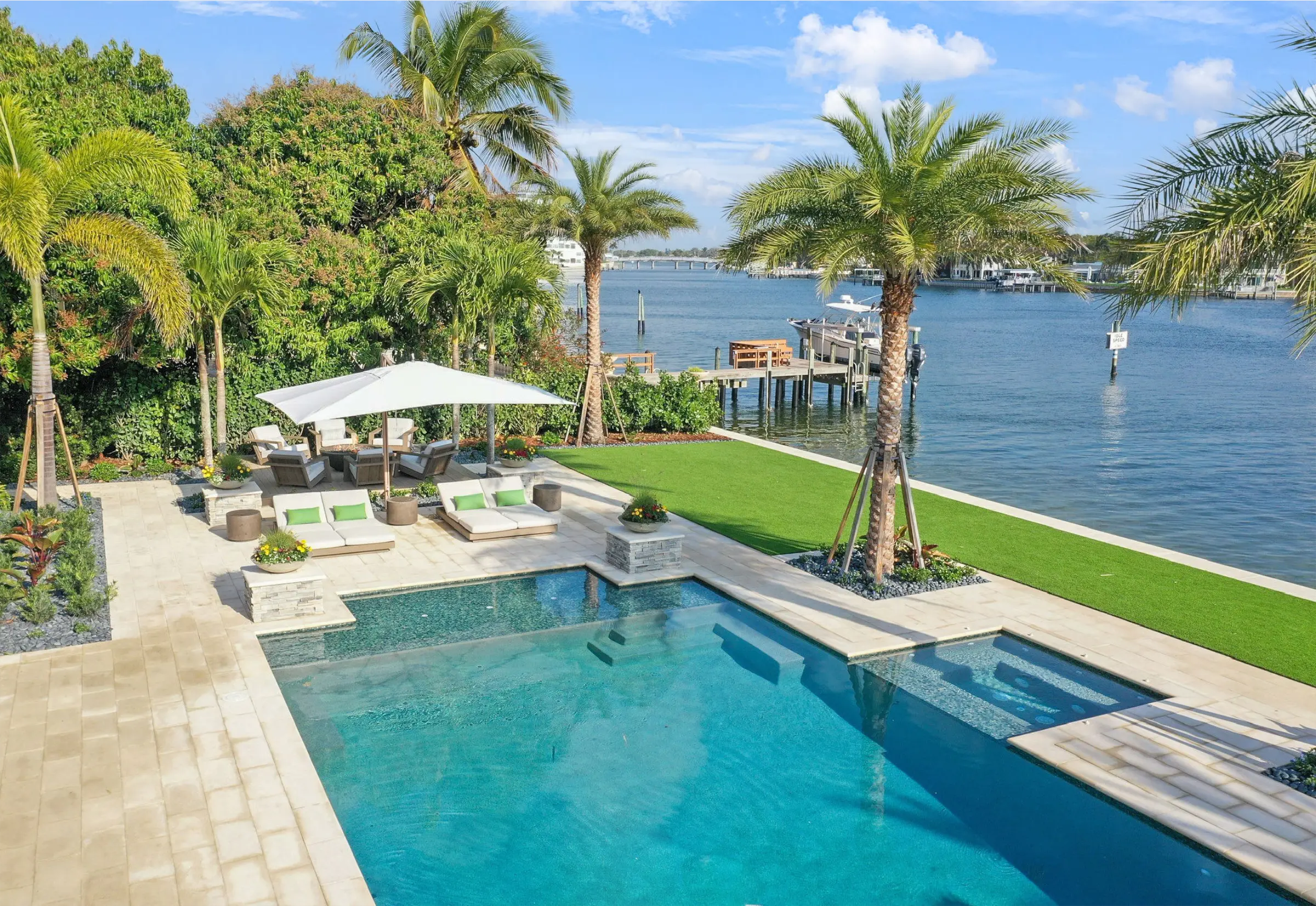 Luxury waterfront pool and spa with manicured lawn, palm trees, and boat dock overlooking St Petersburg bay with residential community in background.
