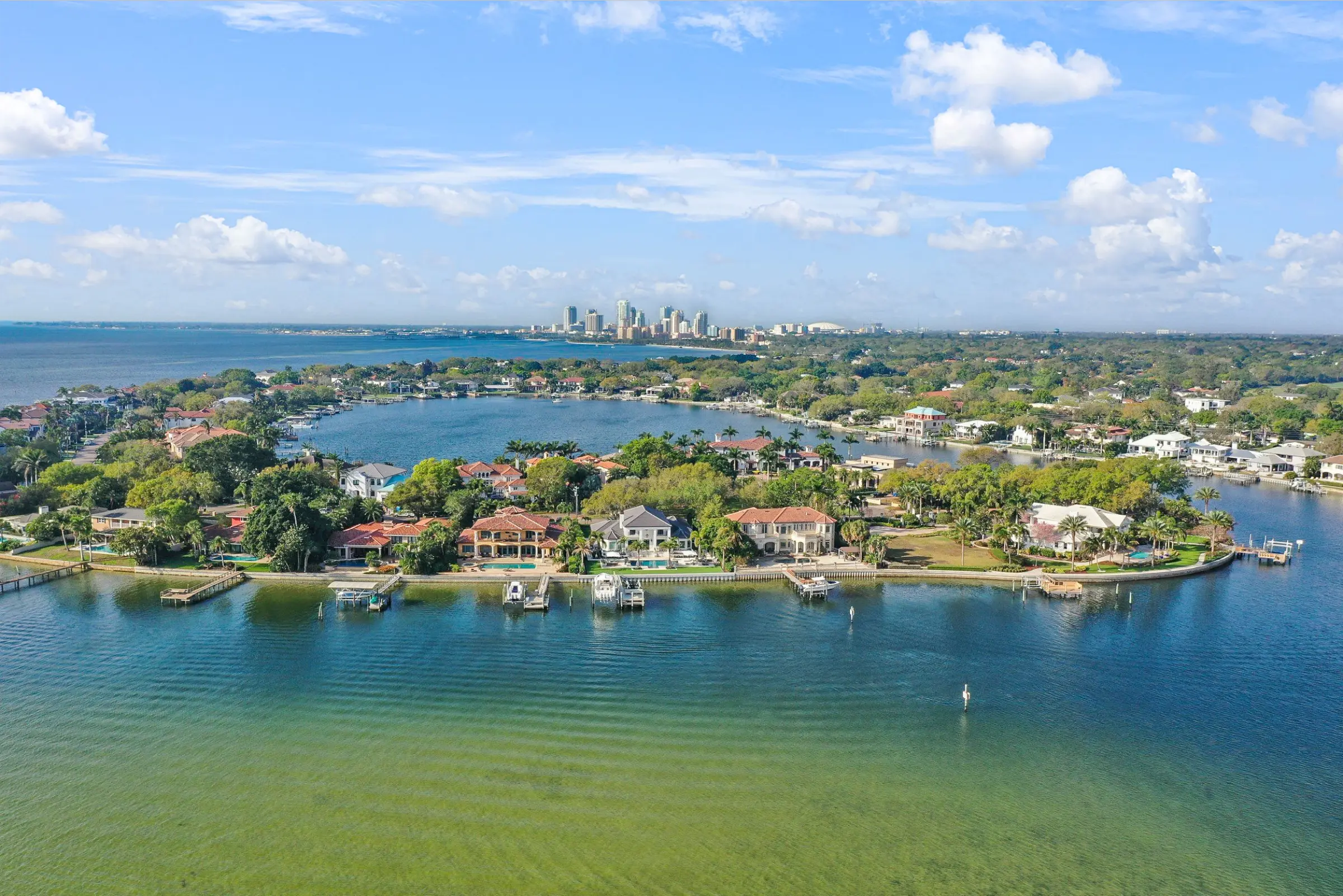 Aerial view of luxury waterfront homes with private docks along Tampa Bay, St Petersburg FL skyline in background across blue water.