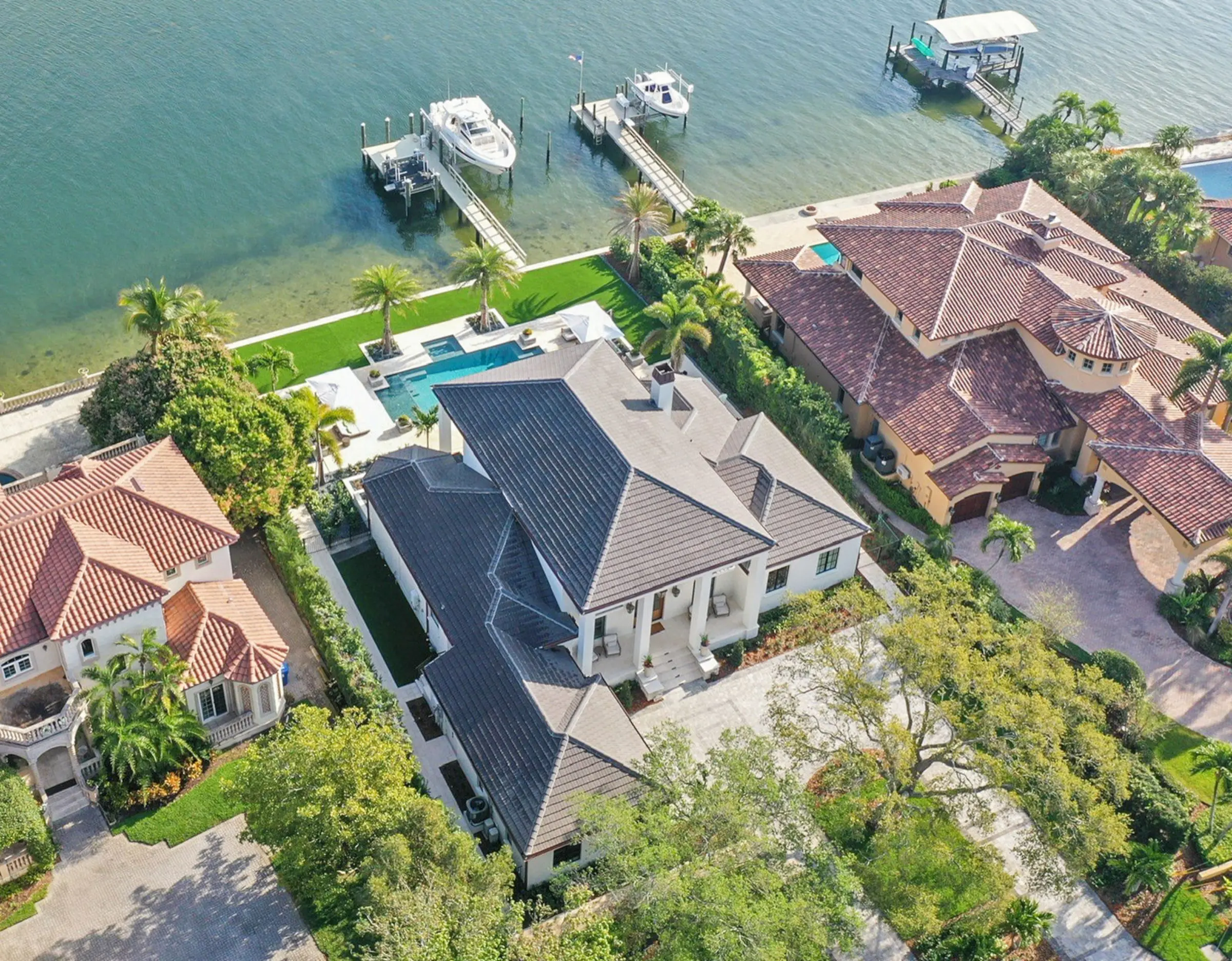 Aerial view of luxury waterfront home with dark metal roof, swimming pool, manicured lawn, and private boat docks on calm turquoise water in St Petersburg, FL.