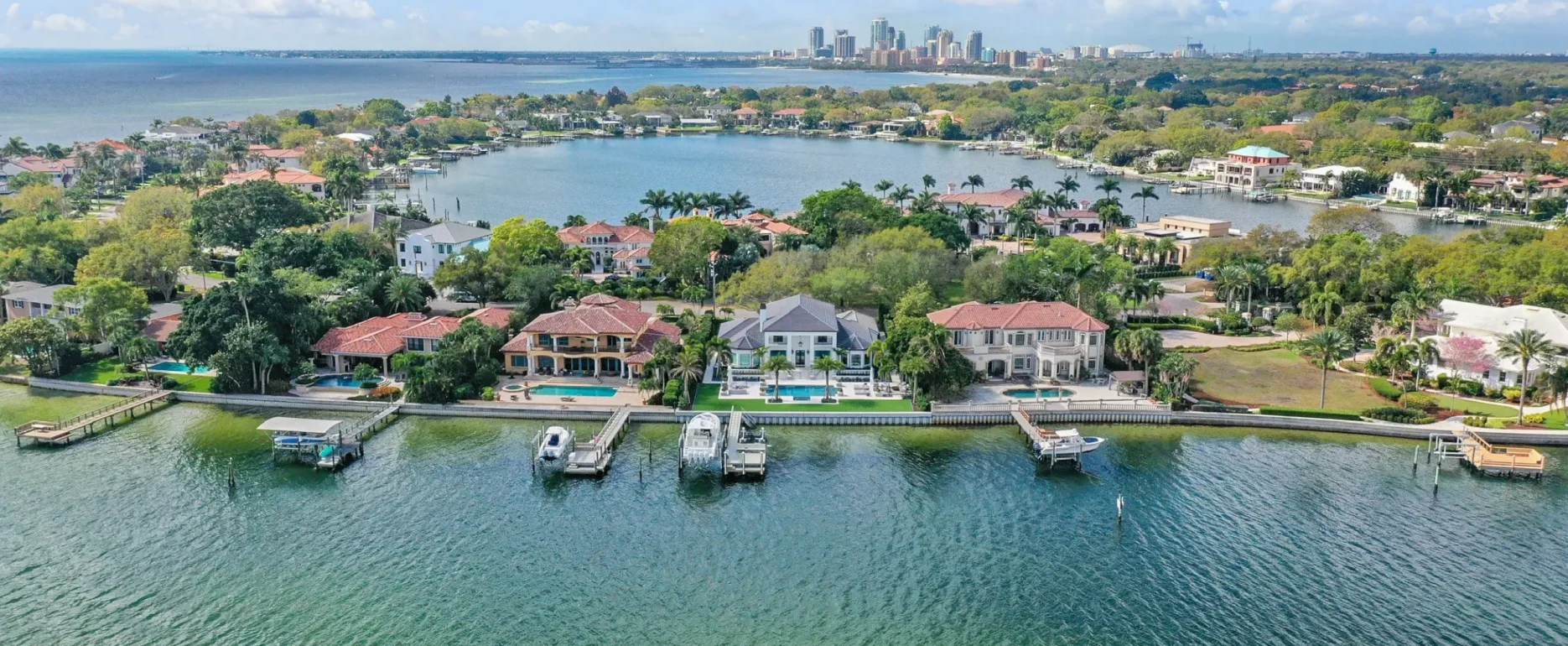 Aerial view of luxury waterfront homes with boat docks along a residential canal, St Petersburg skyline in distance, palm trees and manicured landscaping throughout.