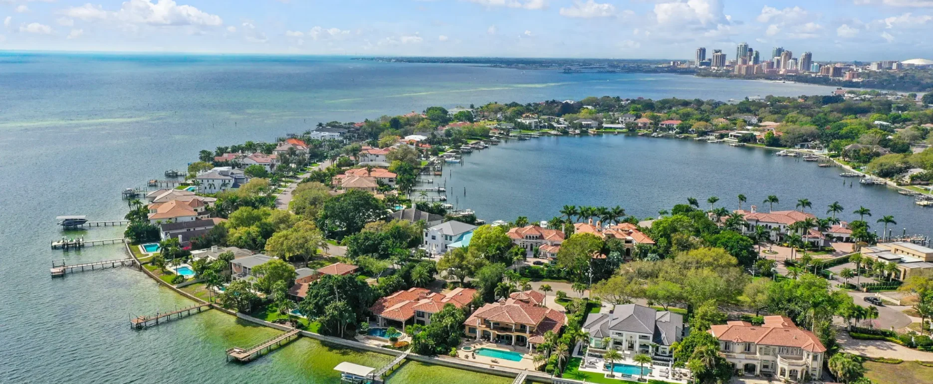 Aerial view of luxury waterfront homes with pools and boat docks along Biscayne Bay, with downtown skyline in distance.