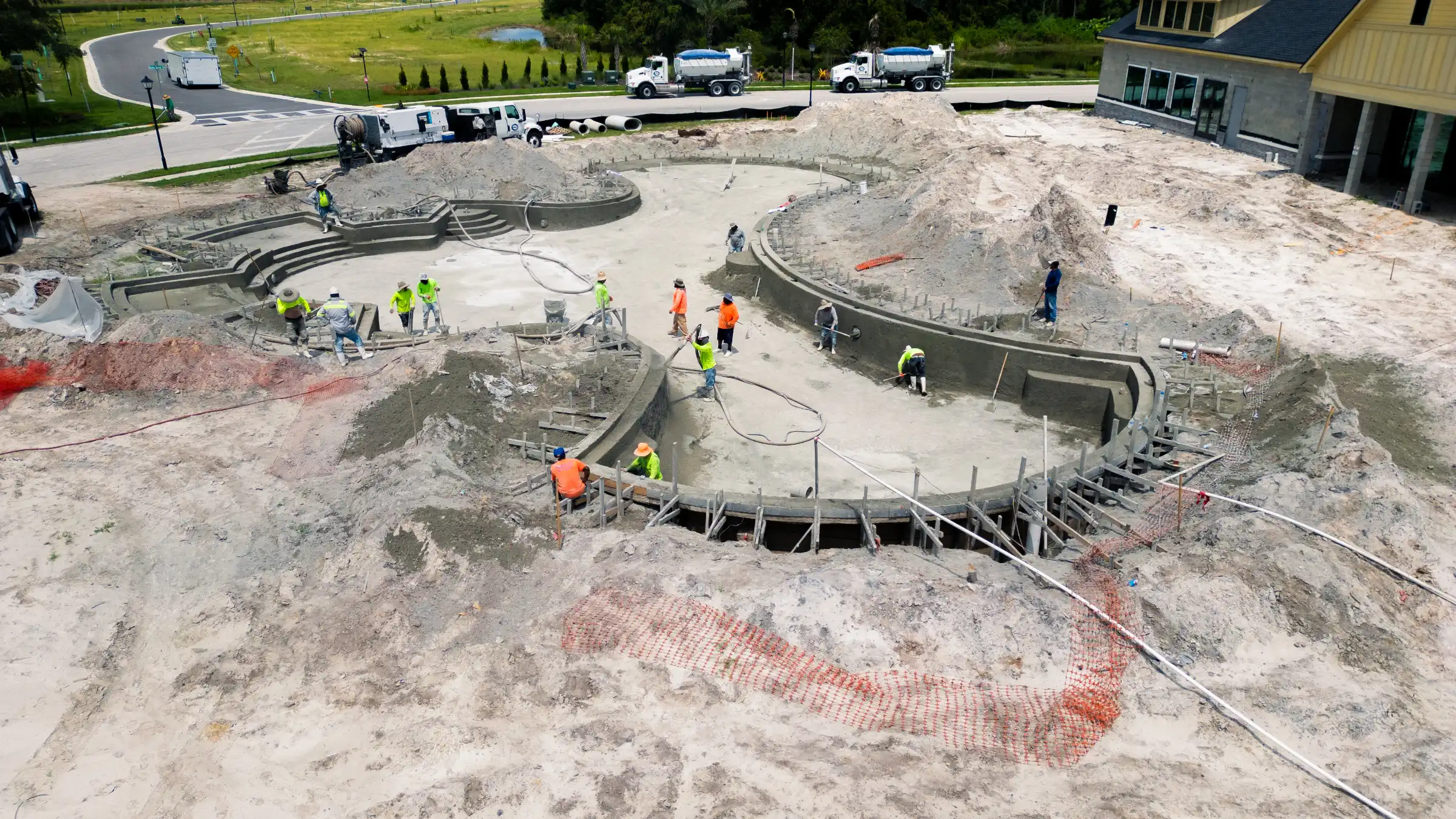 Aerial view of in-progress luxury pool construction in St Petersburg with curved concrete walls, workers in safety gear, and surrounding landscape architecture.