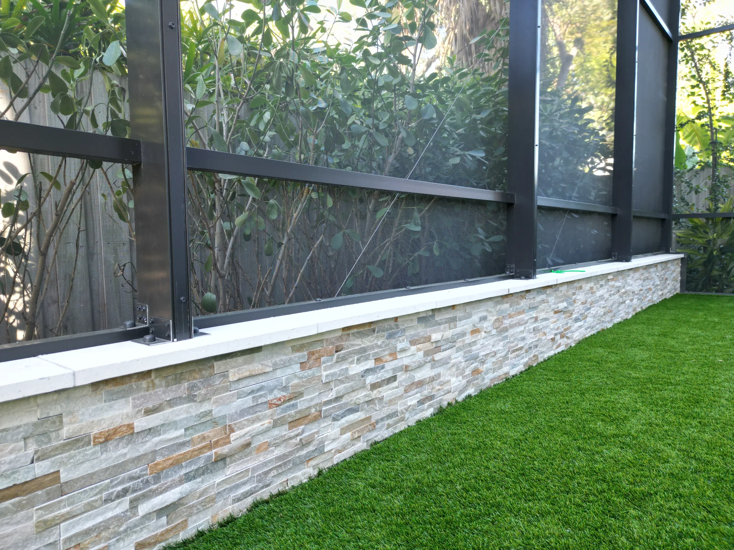 Contemporary black-framed glass structure with stacked stone base and synthetic lawn, exemplifying luxury outdoor construction and landscape architecture.