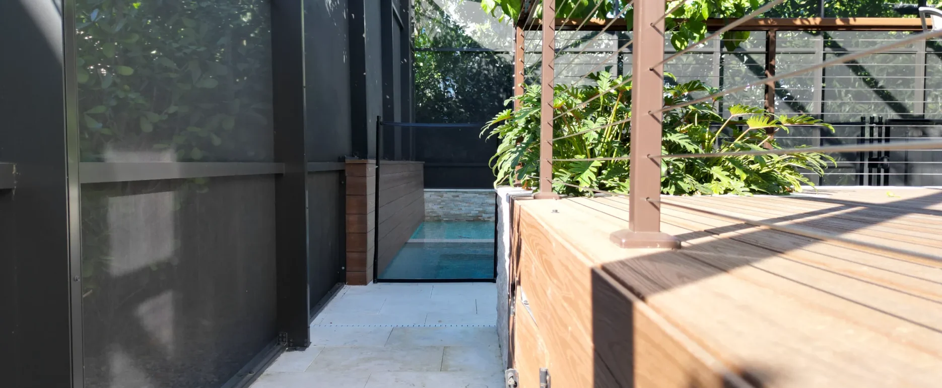 Modern wooden deck with pergola leading to pool area, featuring glass railings and lush climbing vines in luxury residential landscape design.