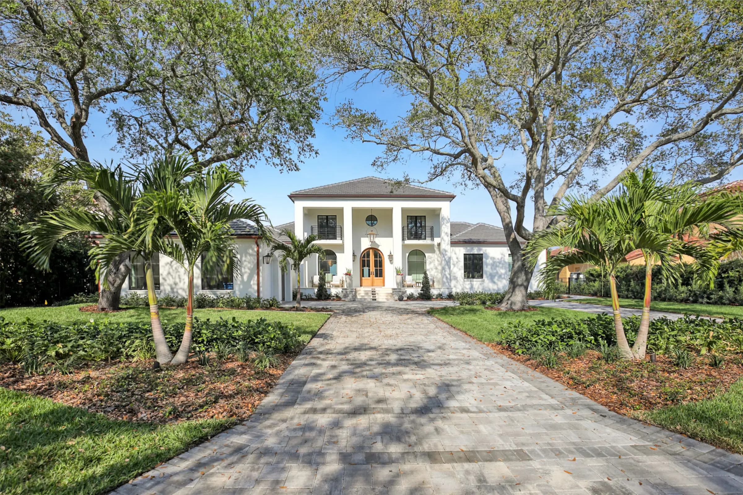 Elegant white two-story colonial home with wooden entry doors framed by manicured palms, brick driveway, and mature shade trees in St Petersburg residential community.