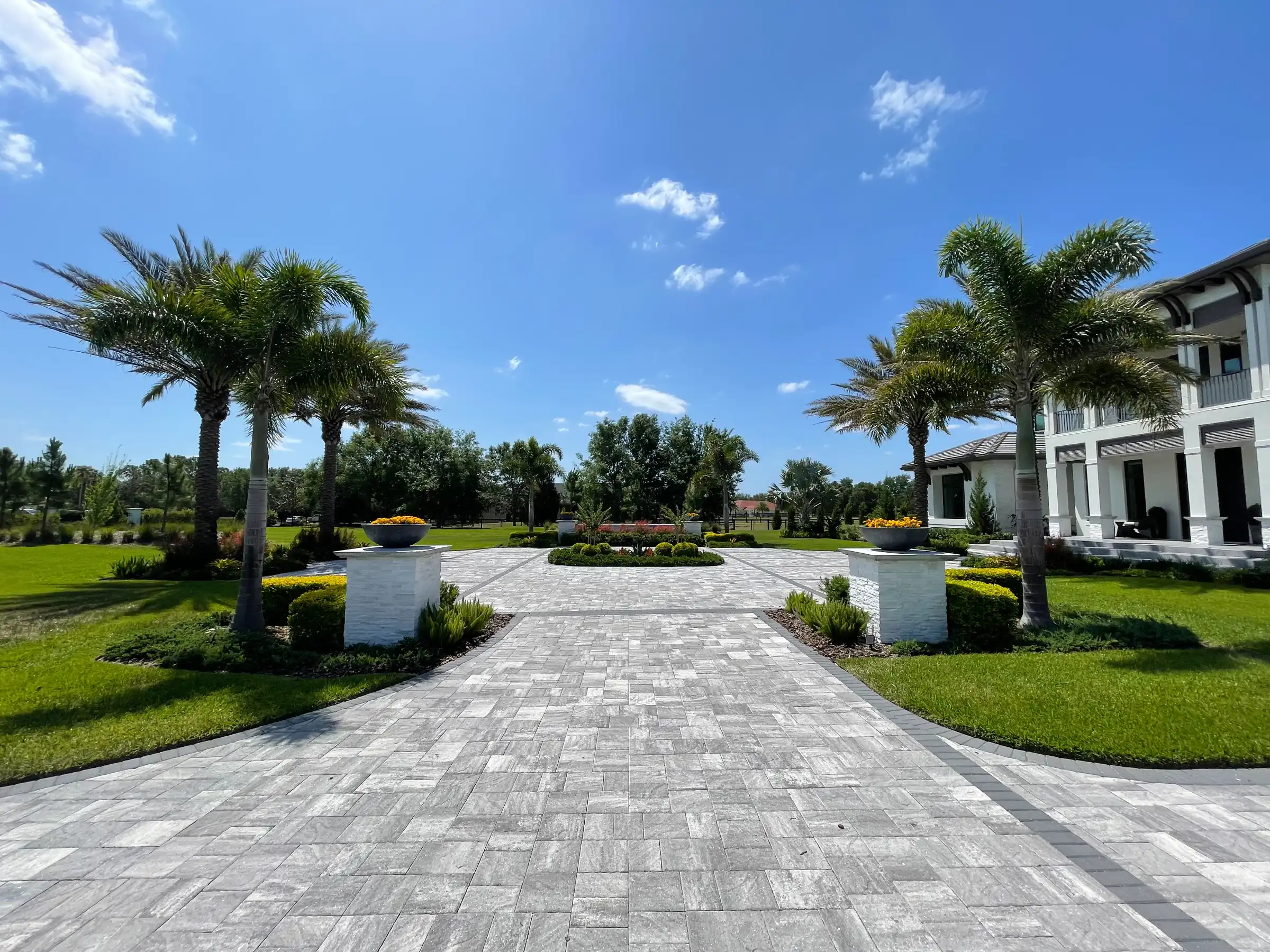 Elegant stone paver driveway lined with tall palms leading to white contemporary home with manicured lawns in luxury residential community.