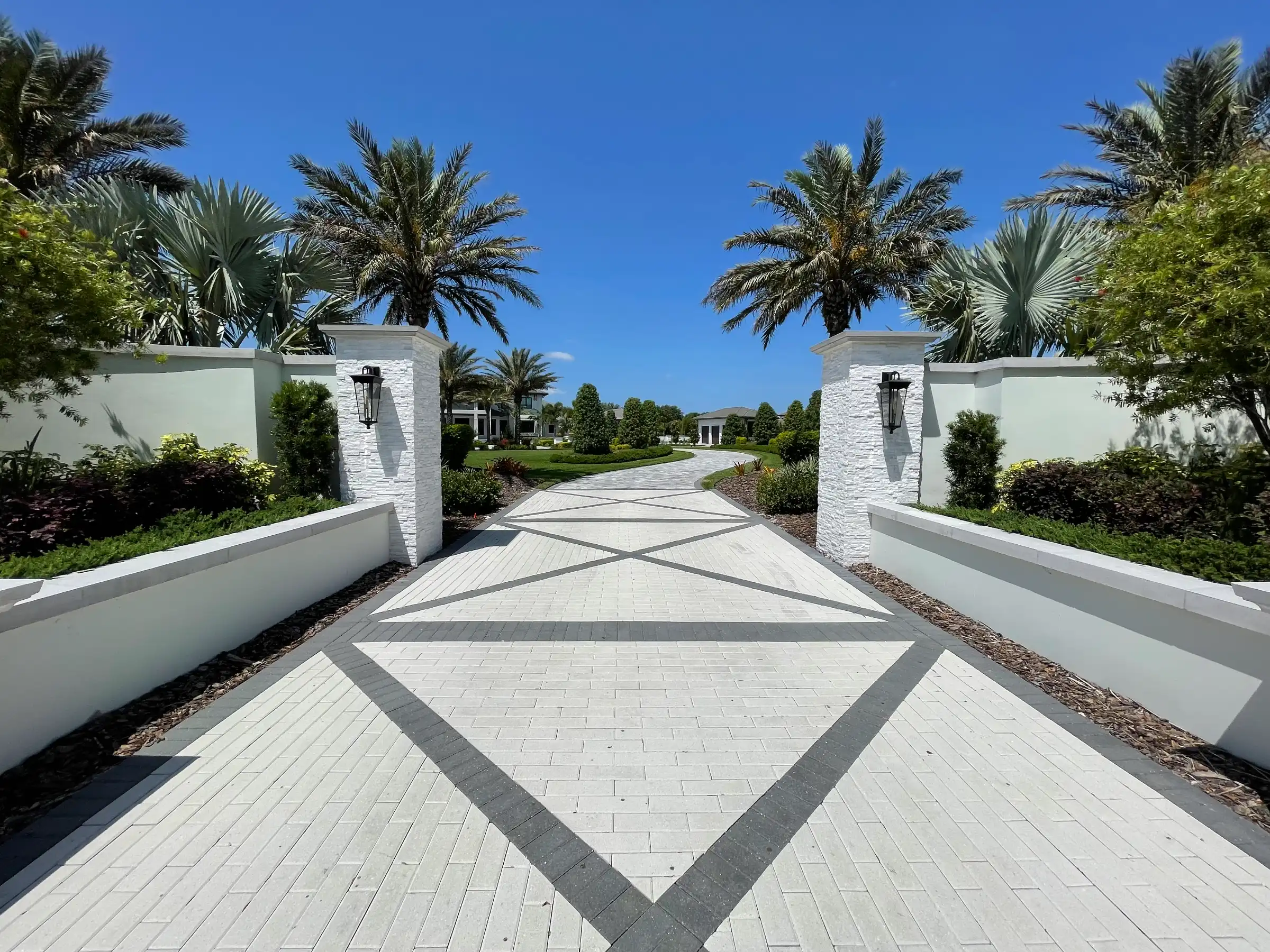 Elegant gated entrance with diamond-patterned pavers leading to luxury residential community in St Petersburg, FL, flanked by white pillars and manicured palms.