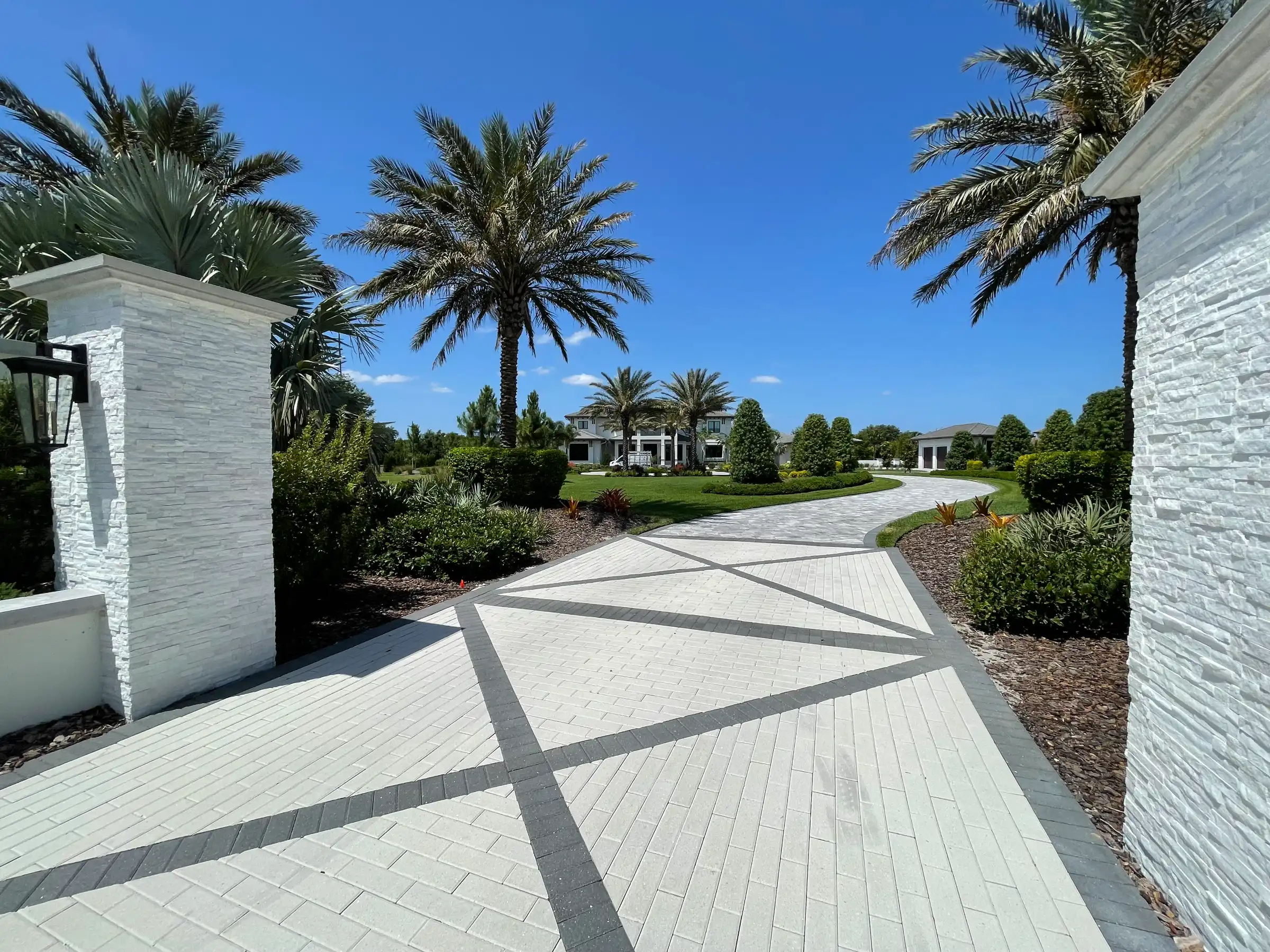 Curved white and gray paver driveway lined with palm trees and manicured landscaping leading to luxury homes in St Petersburg, FL residential community.