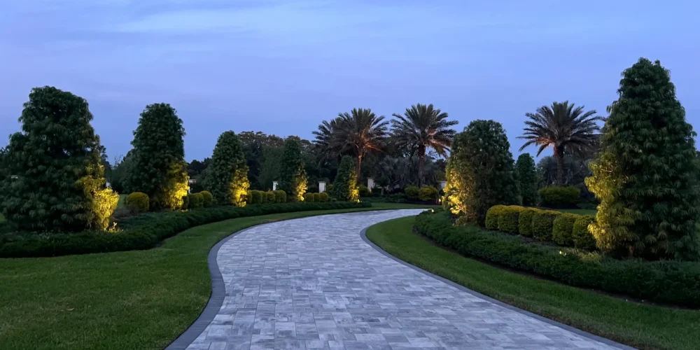 Elegant curved stone paver driveway lined with illuminated trees and manicured hedges at dusk in a luxury St Petersburg residential community.