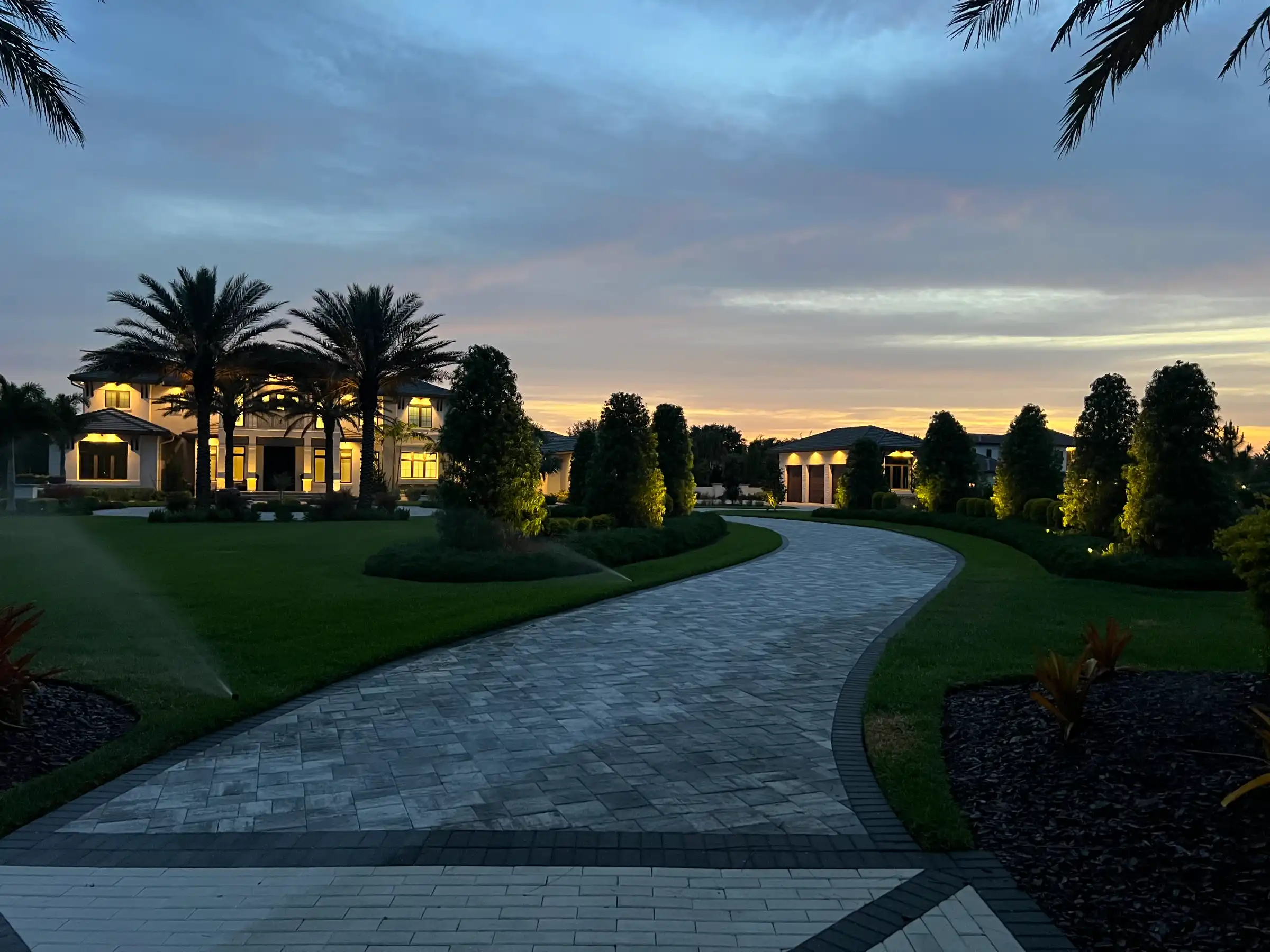 Luxury home at dusk with curved paver driveway, palm trees, and landscape lighting in St Petersburg residential community