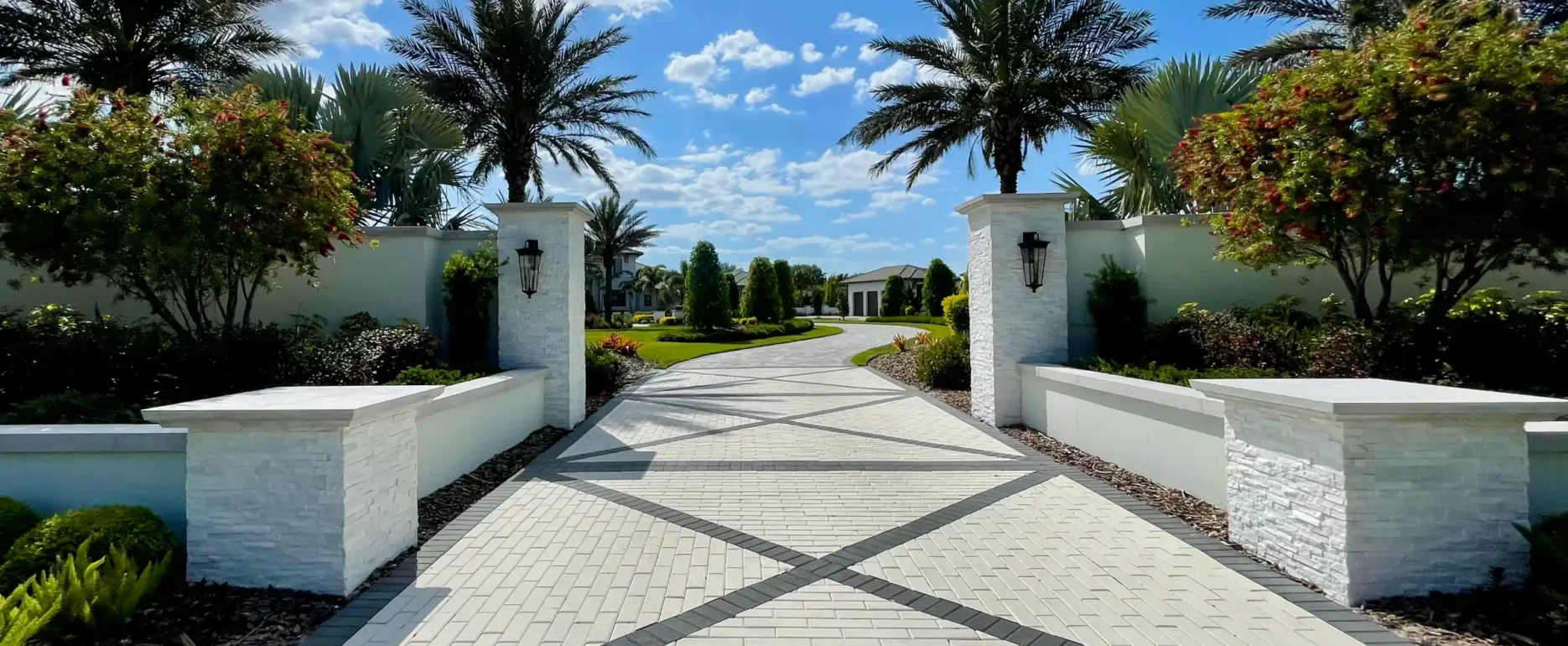 Luxury gated entrance with geometric paver driveway, white stone pillars, and palm-lined landscape design in St Petersburg, FL residential community.