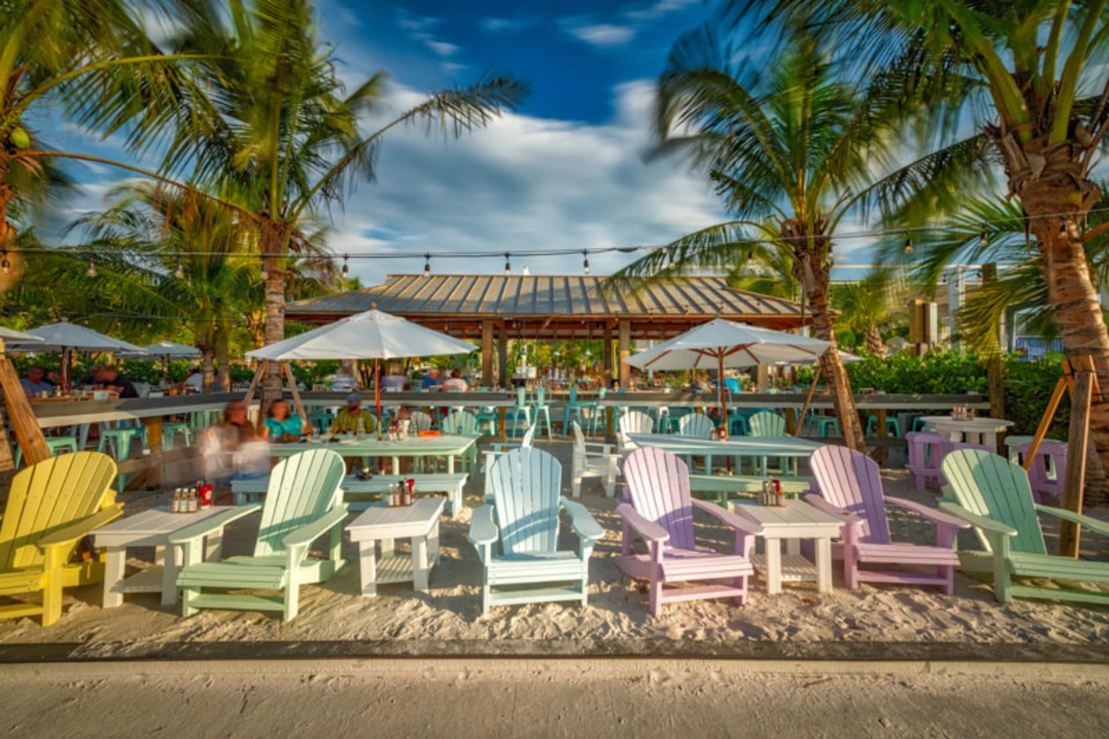 Colorful Adirondack chairs on sandy beach with palm trees, umbrellas, and luxury resort pavilion in St Petersburg FL background.