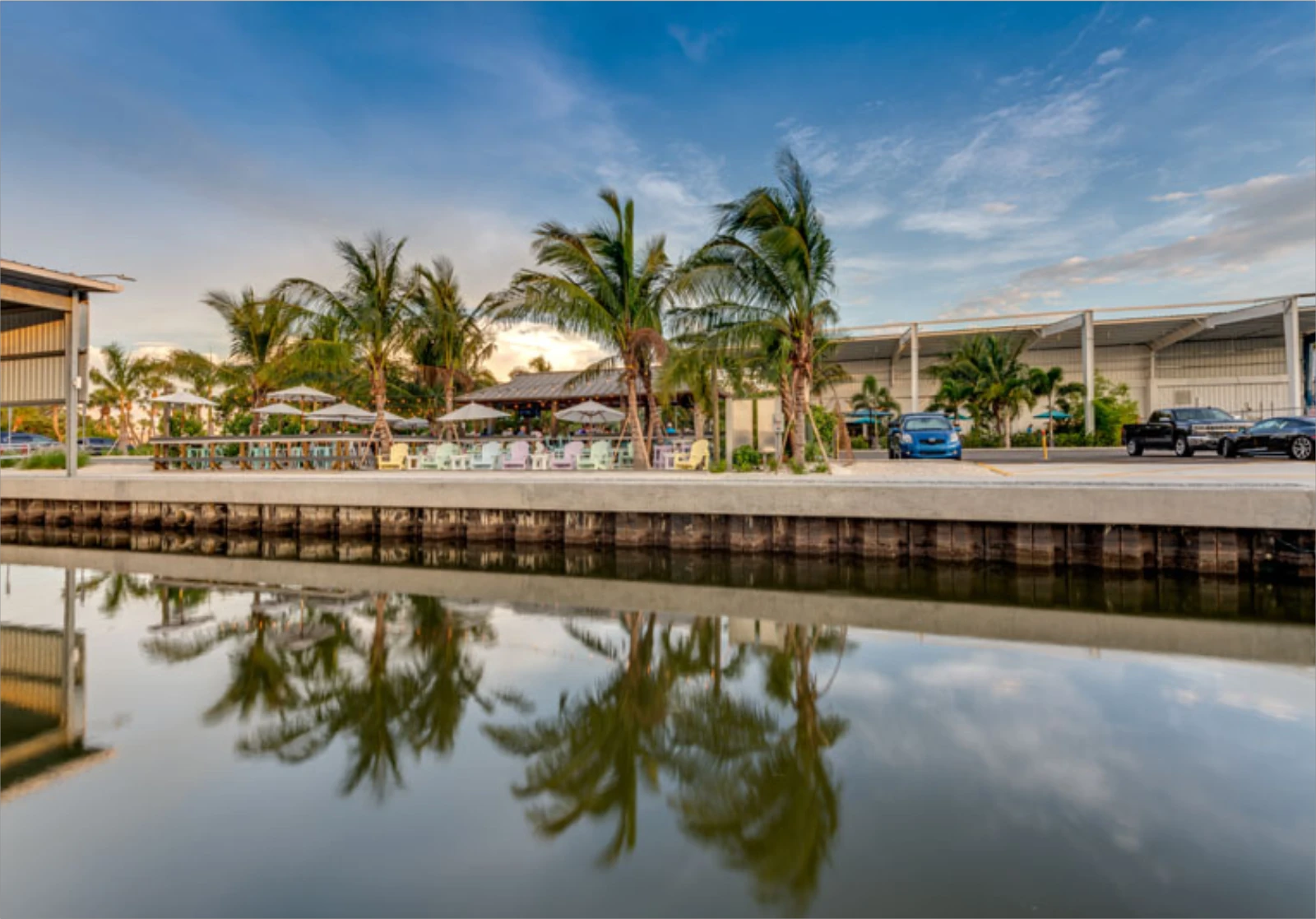 Tranquil waterfront promenade with palm trees, covered seating areas, and parked vehicles reflected in calm water at a St. Petersburg luxury marina development.