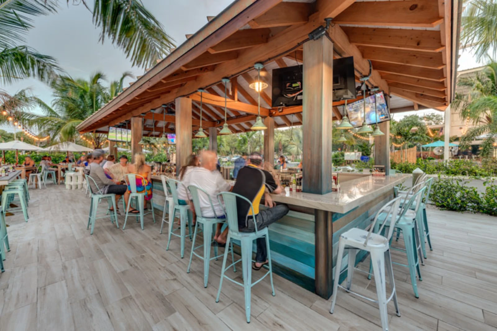 Spacious covered patio with bar seating, wooden beams, light blue metal chairs, and palm tree landscaping at a St Petersburg coastal venue.