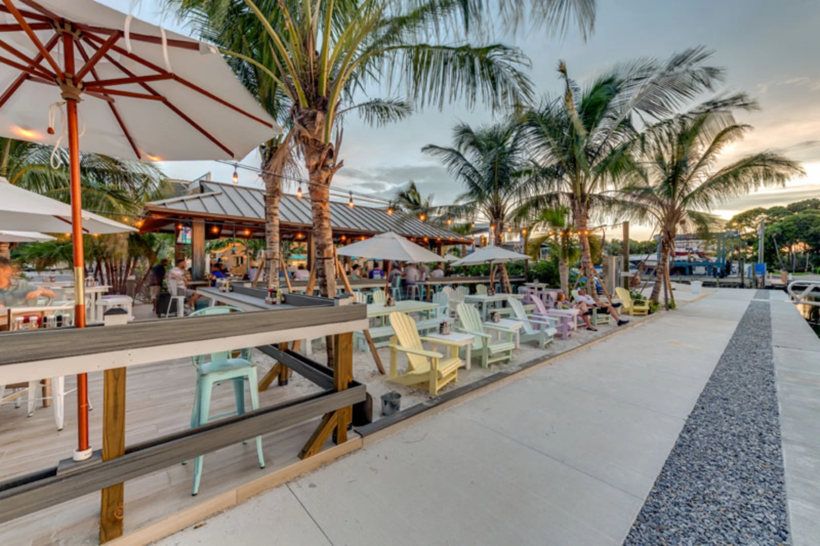 Modern waterfront dining area with pastel Adirondack chairs, wooden deck, palm trees, and covered pavilions at a luxury St Petersburg Florida resort.