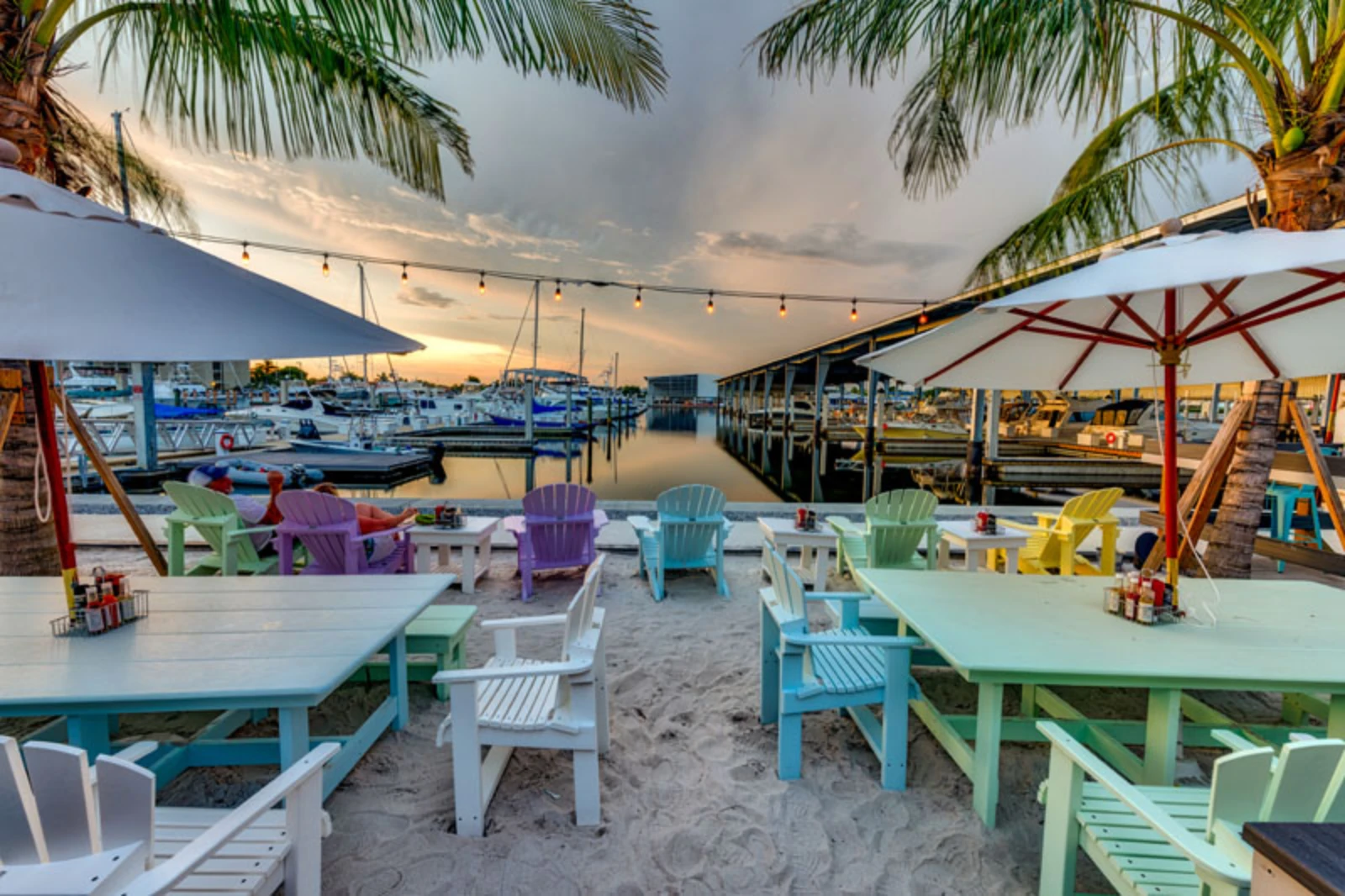 Vibrant waterfront dining area with multicolored Adirondack chairs, tables, and umbrellas overlooking a marina at sunset in St Petersburg, FL.