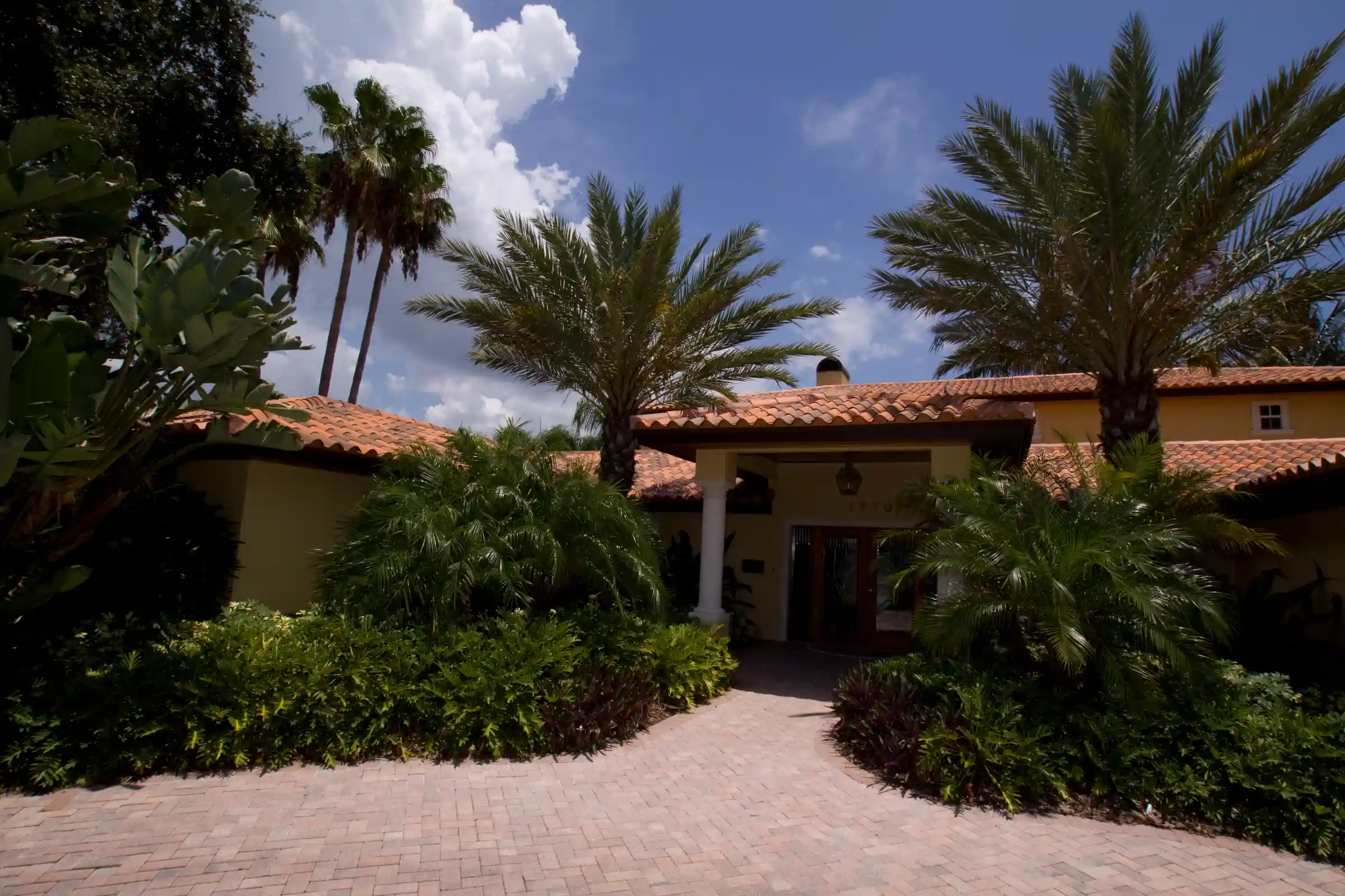 Spanish colonial residence with terracotta tile roof, cream stucco walls, and mature palm trees lining brick courtyard entrance in St Petersburg luxury community.